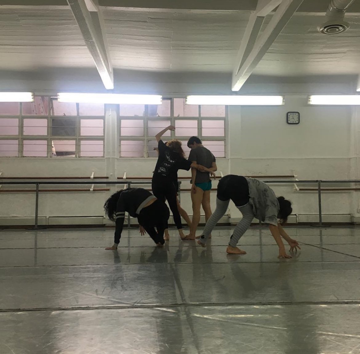 Five dancers rehearsing in a dance studio with mirrored walls, ballet barres, overhead lighting, and a clock on the wall.