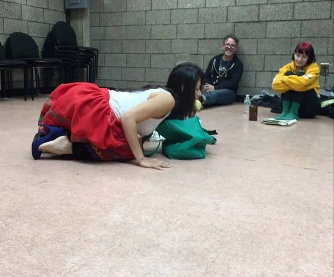 A woman kneeling on the floor, leaning forward with her face close to a green bag, in a room with a brick wall. Two other people are sitting on the floor in the background, one smiling and the other with arms crossed.