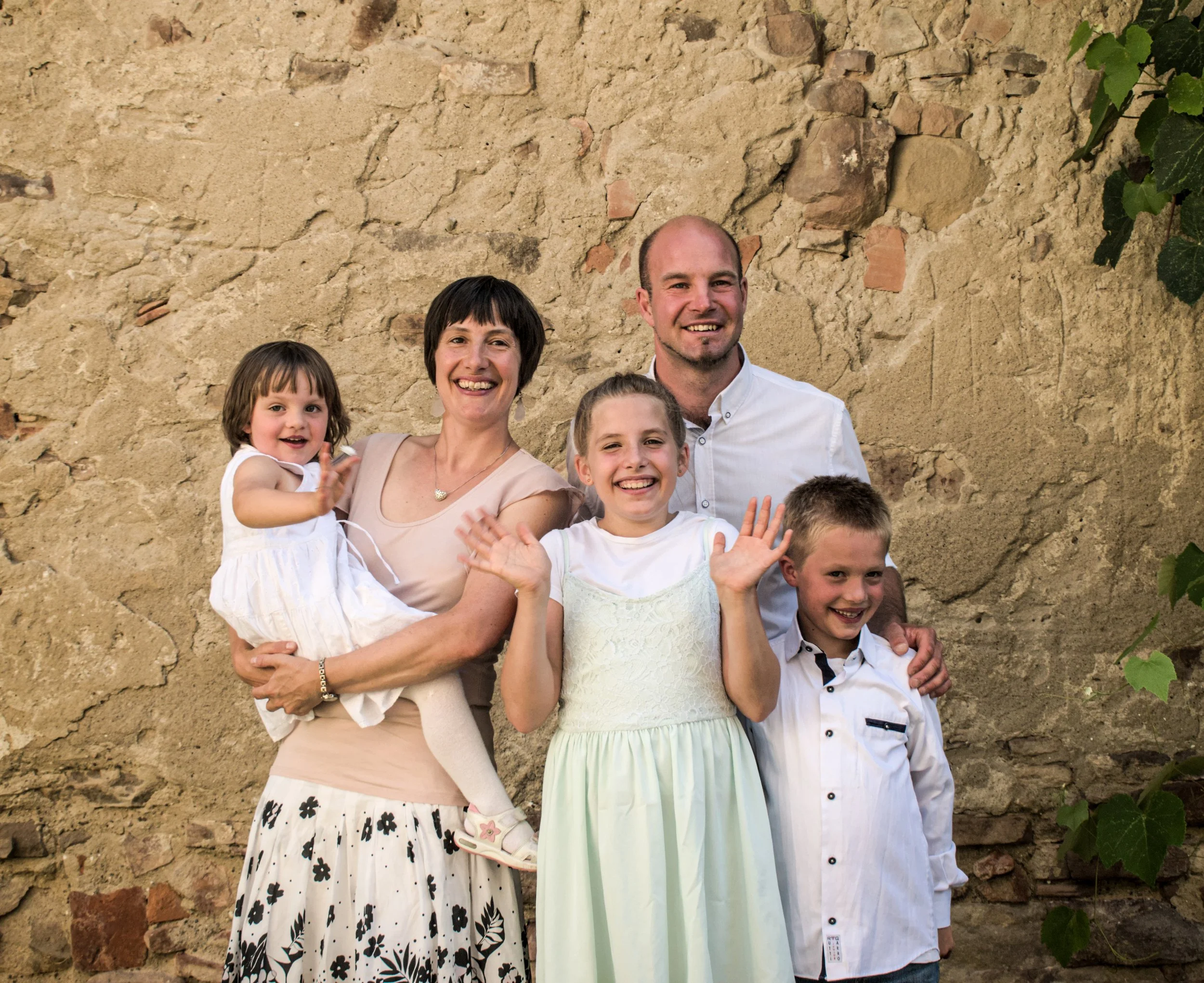 Familie mit zwei Erwachsenen und drei Kindern steht vor einer alten Steinmauer, alle lächeln und winken in die Kamera.