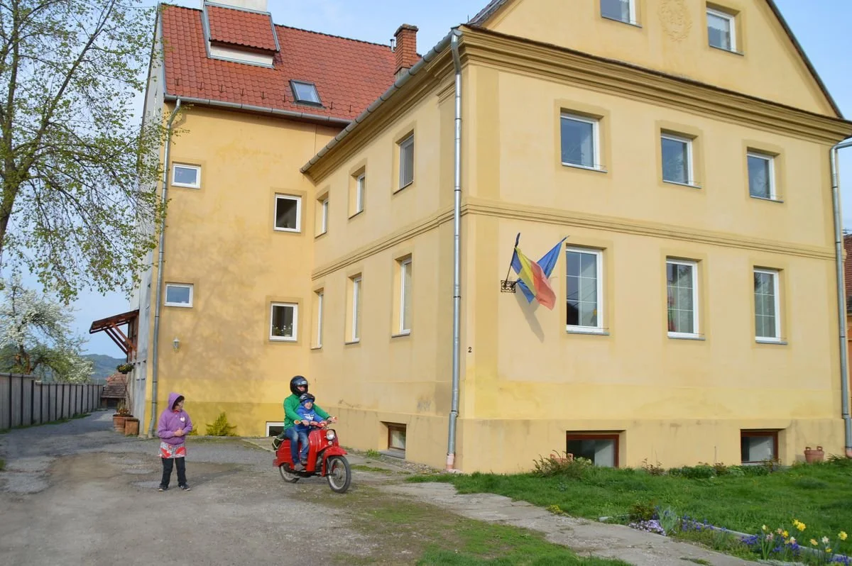 Ein gelbes Mehrfamilienhaus mit sieben Fenstern und drei Dachfenstern, eine Flagge an der Wand, zwei Kinder im Gespräch, eines auf einem roten Motorroller mit Helm, das andere steht daneben in einem lila Jacke, im Vorgarten blühende Blumen.