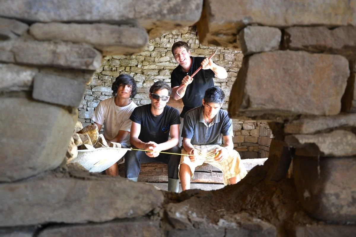 Vier Menschen in einer alten, steinernen Struktur, eine Person hält einen Hammer, alle schauen durch ein Loch in der Wand, die Szene wirkt wie bei einer archäologischen Ausgrabung.