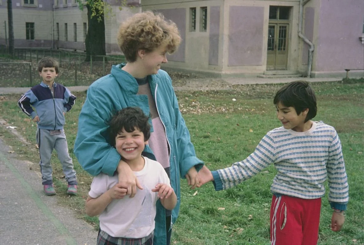 Eine Frau und zwei Jungen in einem Park spielen und lachen, während ein dritter Junge im Hintergrund zusieht.