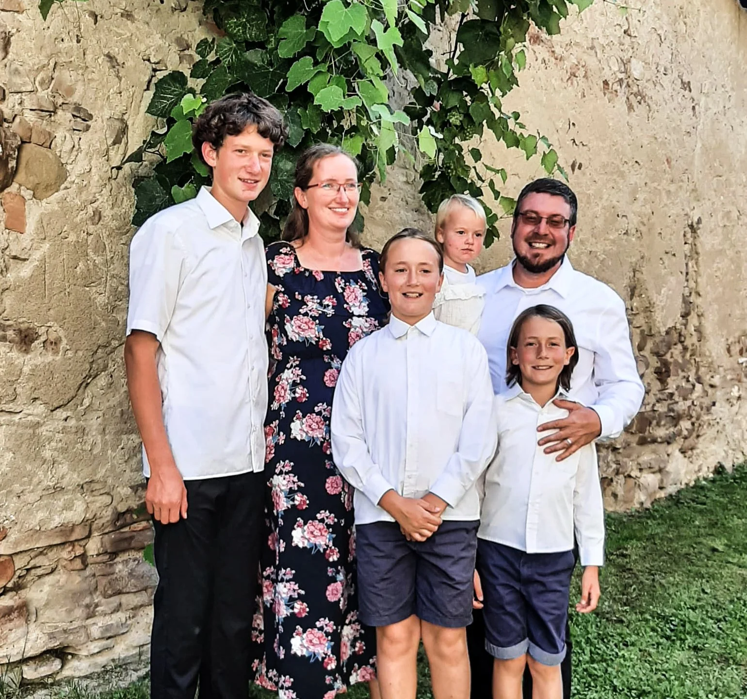 Eine Familiengruppe bestehend aus fünf Erwachsenen und zwei Kindern, steht vor einer alten Steinmauer mit grünen Pflanzen. Sie sind alle freundlich lächelnd, gekleidet in weiße Hemden und dunkle Hosen oder Röcke.