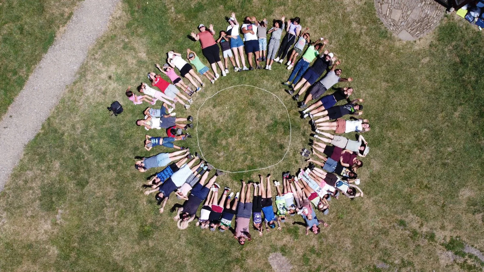 Blick von oben auf eine Gruppe von Menschen, die in einem Kreis auf einer Wiese liegen. Sie sind in einem Außengelände, möglicherweise in einem Park, mit einer kleinen gepflasterten Fläche im Hintergrund.