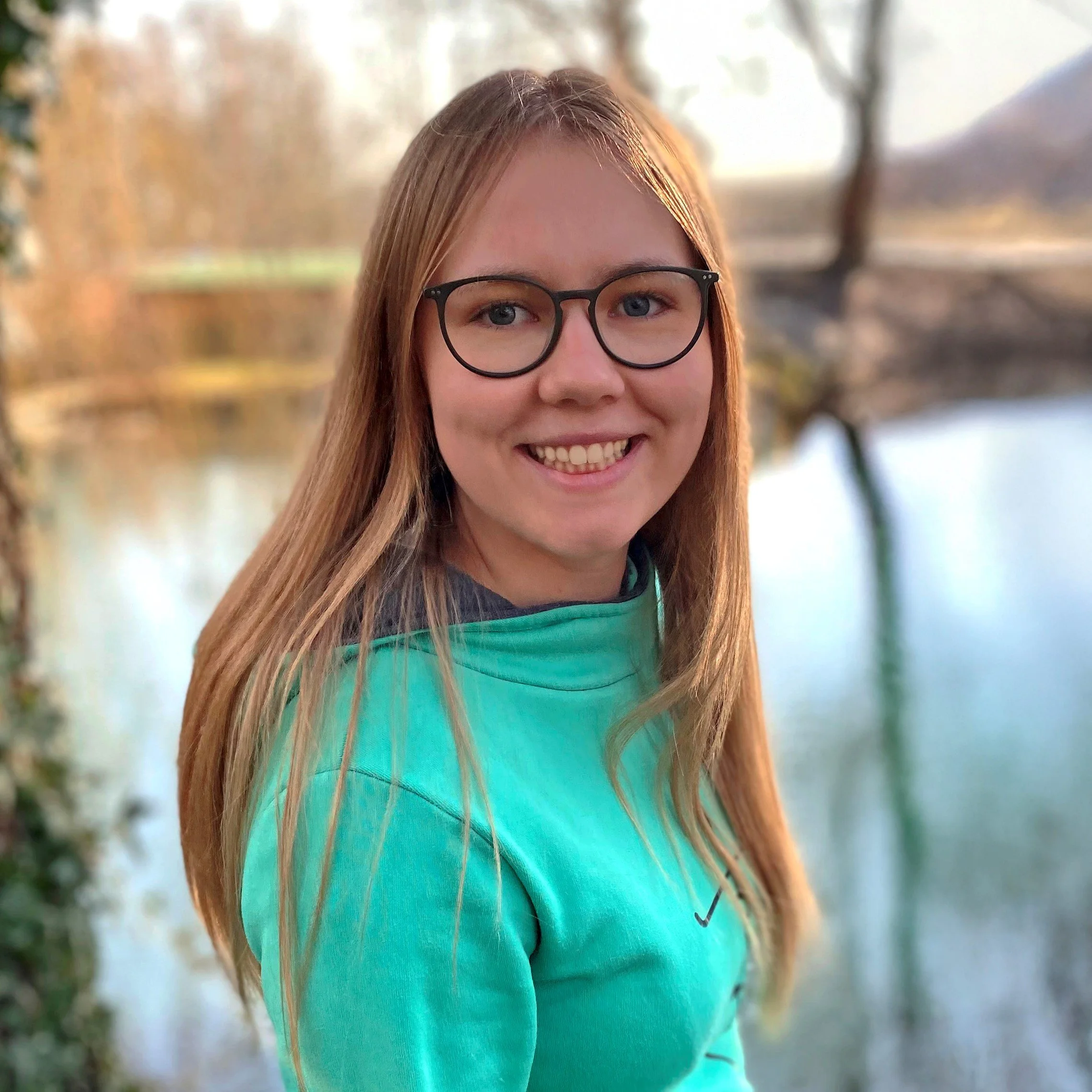 Junge Frau mit Brille und braunen Haaren, lachend, im Freien an einem Fluss, trägt eine türkise Jacke.