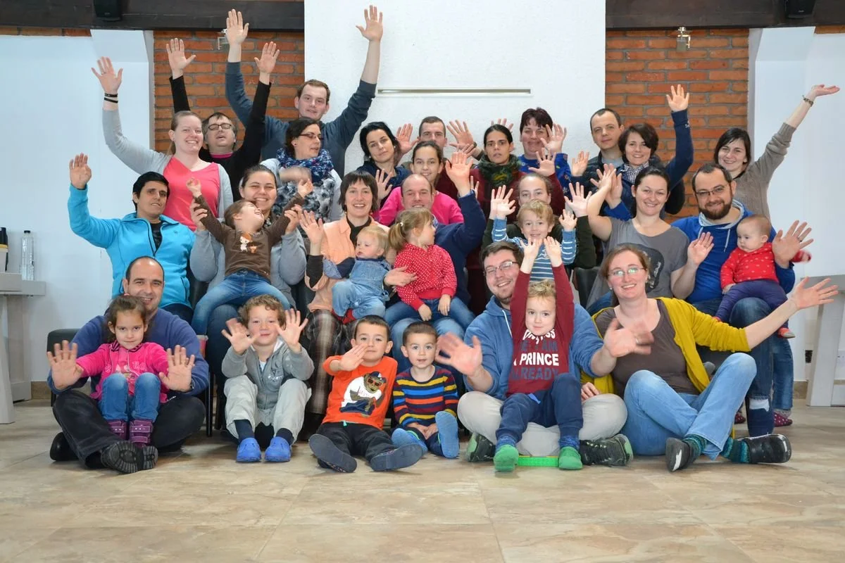 Gruppenfoto von Menschen unterschiedlichen Alters, die im Raum vor einer weißen Wand und einer roten Backsteinwand sitzen und stehen, alle winken und lächeln.