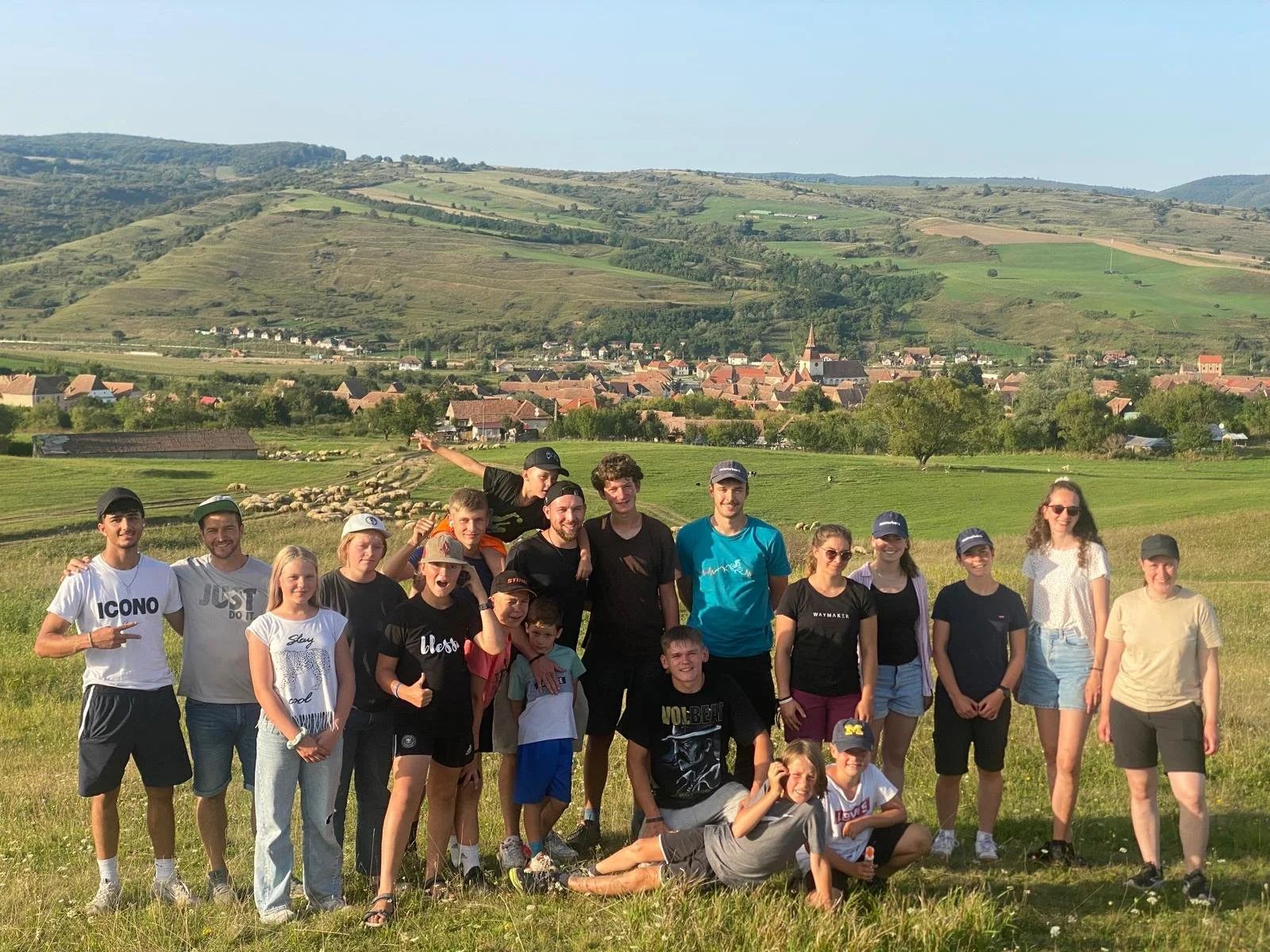 Gruppe von jungen Menschen in einem grünen Feld mit Hügeln und einem kleinen Dorf im Hintergrund.