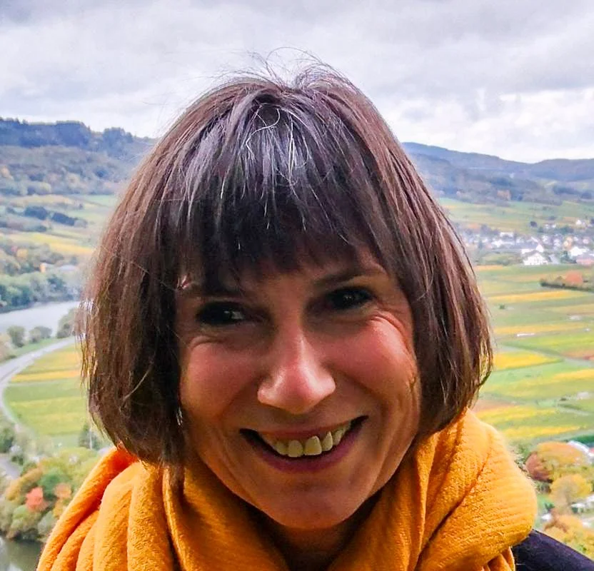 Lächelnde Frau mit braunem Haar vor einer ländlichen Landschaft mit Fluss und Hügeln im Hintergrund.
