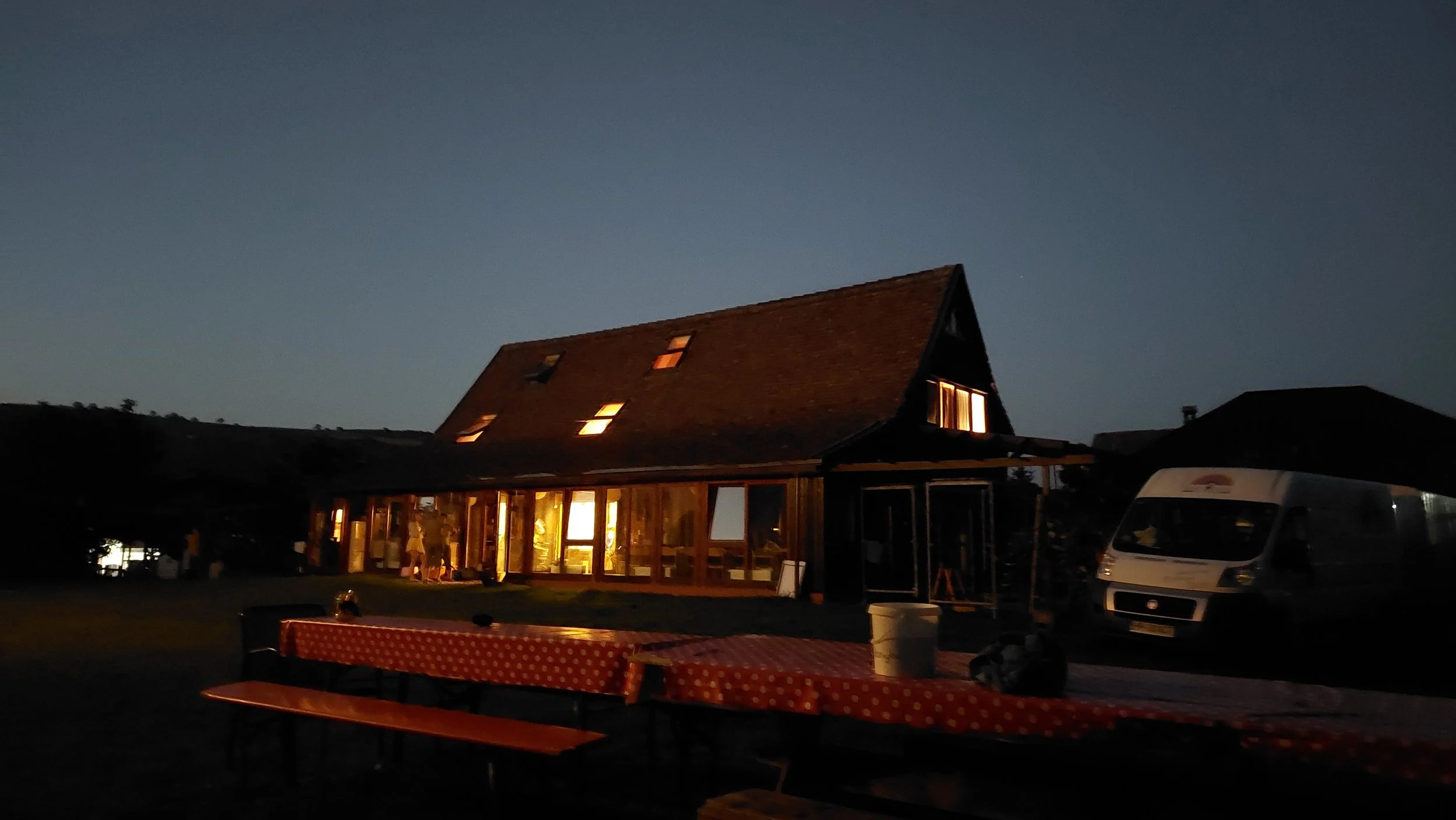 Ein Haus mit großen Fenstern, das nachts warm beleuchtet ist, sowie eine Motorhaube mit einem roten Tisch im Vordergrund bei dunklem Himmel.