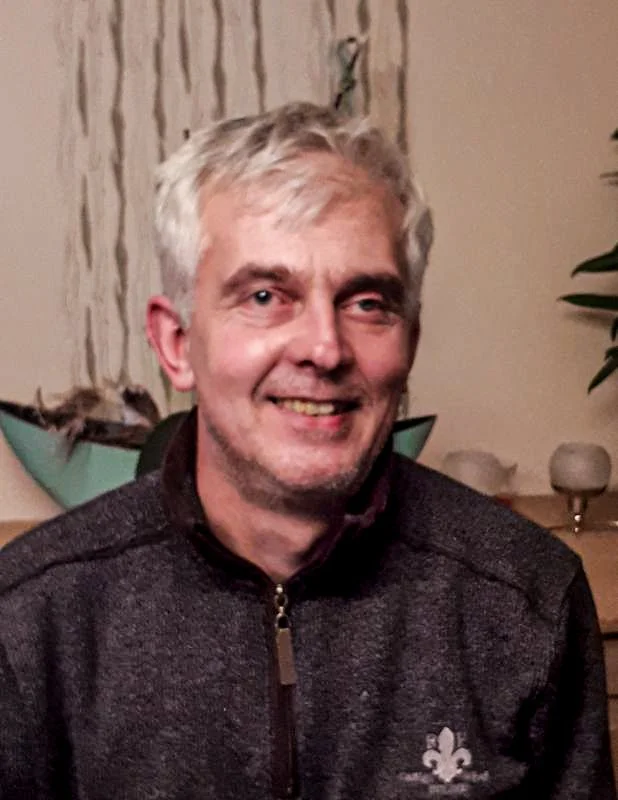 Ein lachender Mann mit grauem Haar und dunkler Jacke, sitzend in einem Raum mit Dekorationen im Hintergrund.