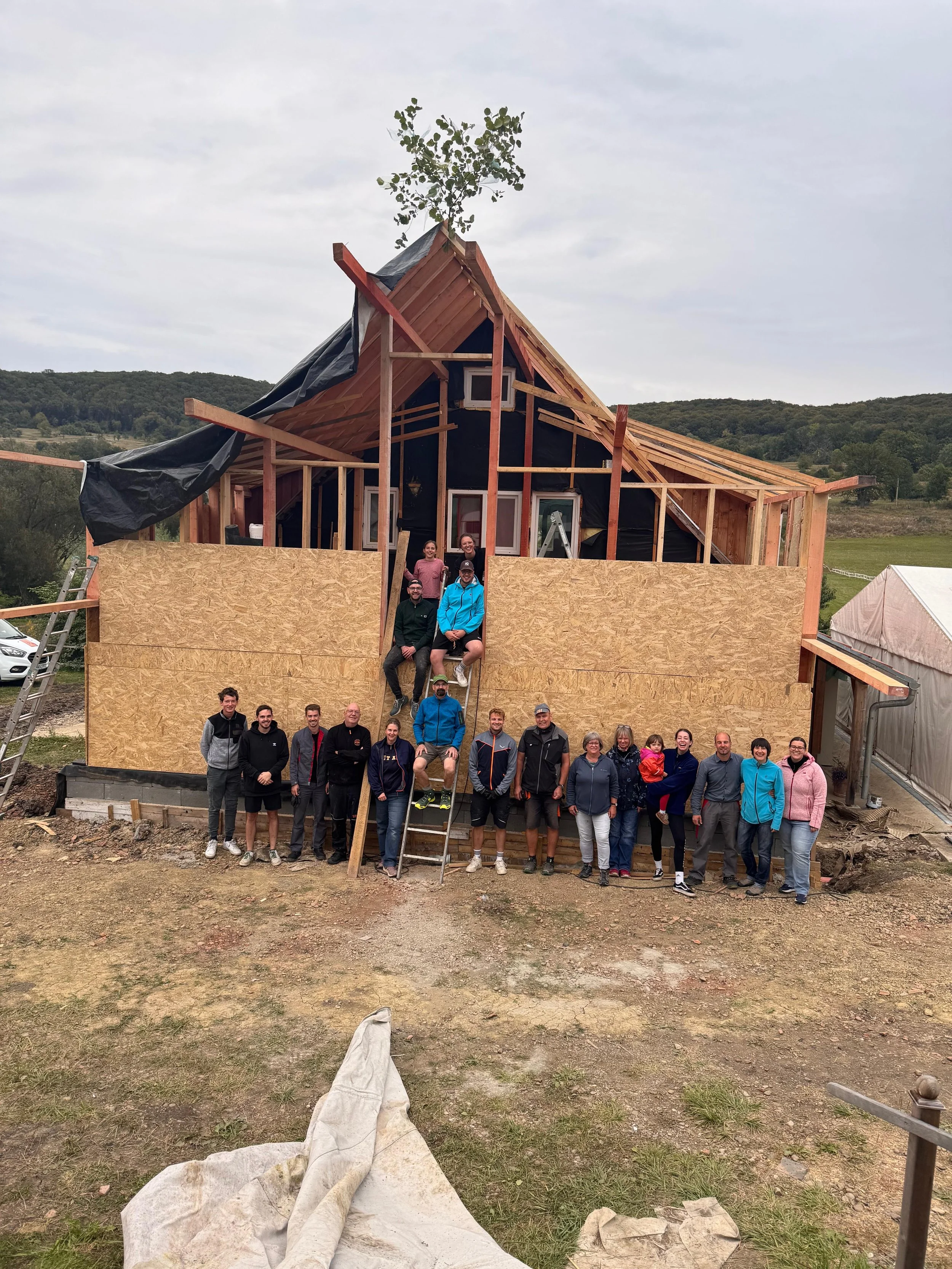 Gruppe von Menschen vor einem im Bau befindlichen Haus, das überwiegend aus Holzrahmen und Sperrholzplatten besteht, im Freien mit einer ländlichen Landschaft im Hintergrund.