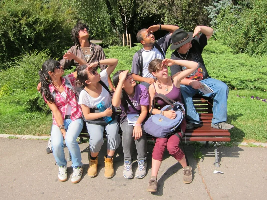 Eine Gruppe von sieben jungen Menschen sitzt auf einer Parkbank im Freien, einige schauen in die Sonne, während andere in verschiedene Richtungen blicken, um den Himmel zu beobachten.