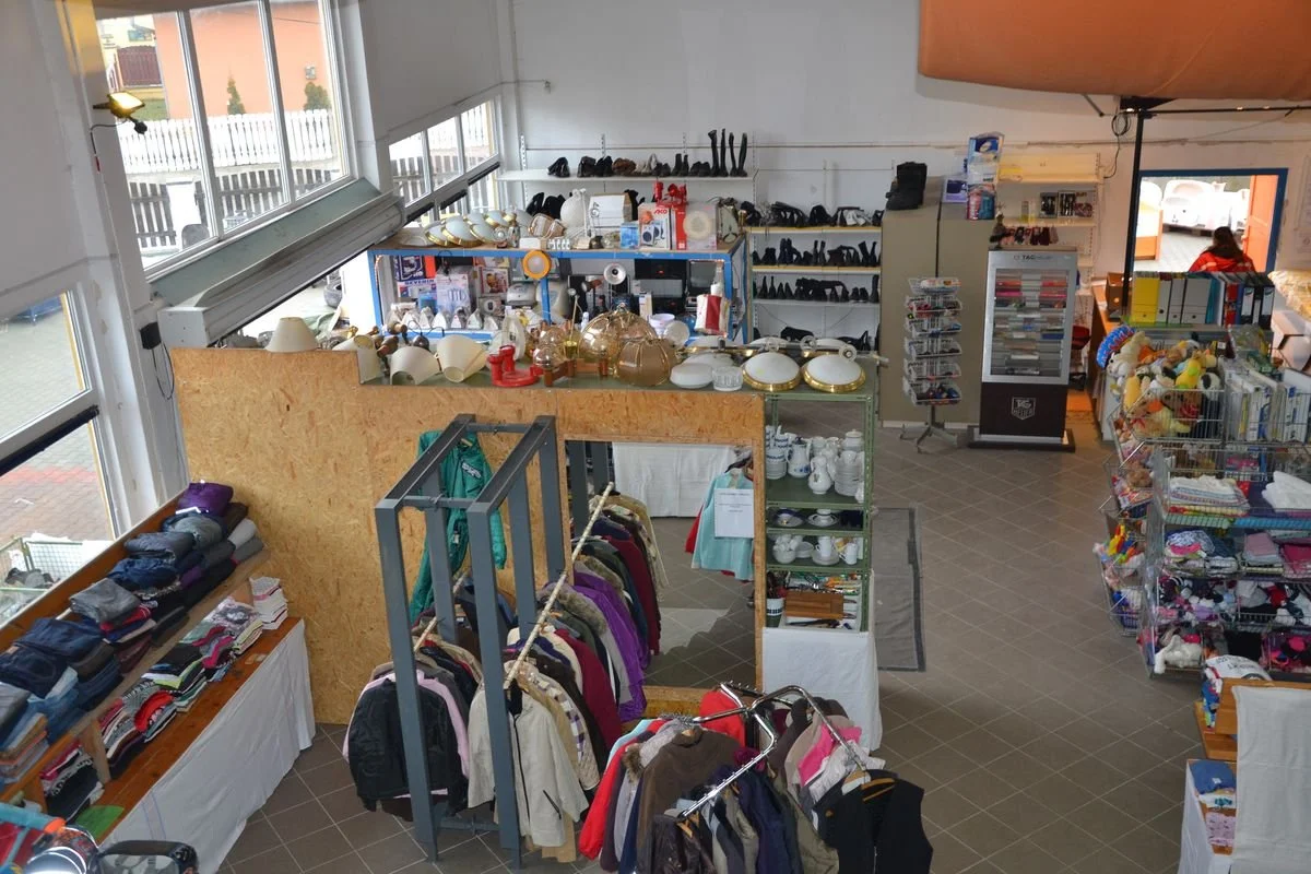 Ein Second-Hand-Laden mit Kleidung, Geschirr, Schuhen und Spielzeug in Regalen und auf Tischen, gut beleuchtet mit großen Fenstern.