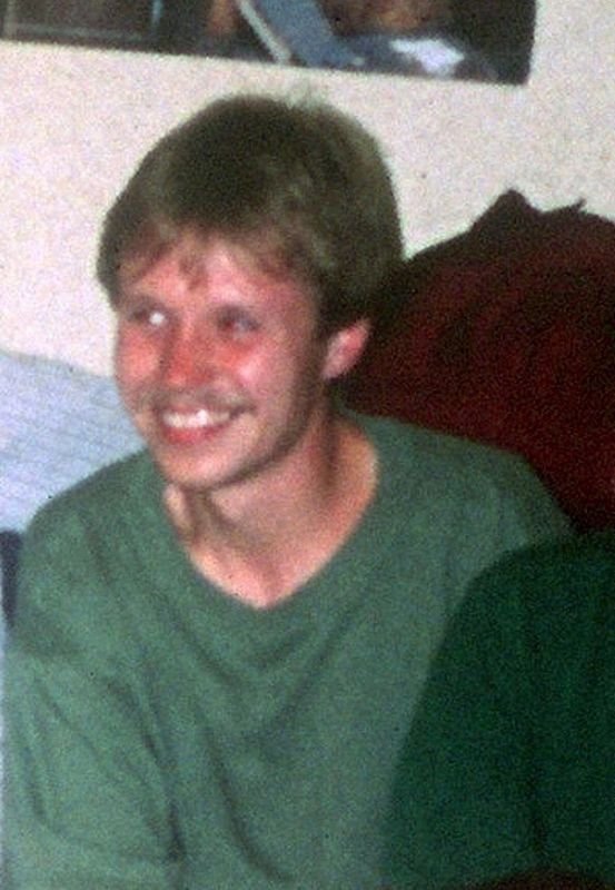 Ein junger Mann mit braunen Haaren, grünem T-Shirt und einem freundlichen Lächeln sitzt in einem Raum.