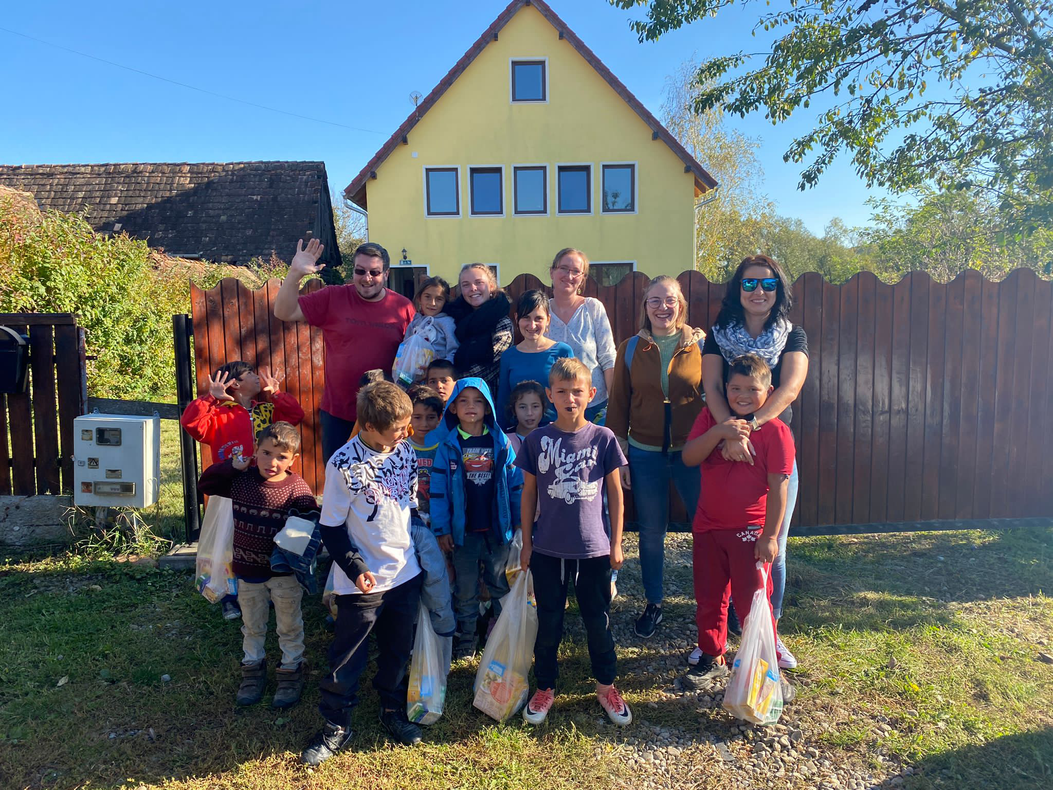 Gruppe von Erwachsenen und Kindern vor einer Hecke, einige Kinder halten Einkaufstüten. Es ist ein sonniger Tag, im Hintergrund ein gelbes Haus mit rotem Dach.
