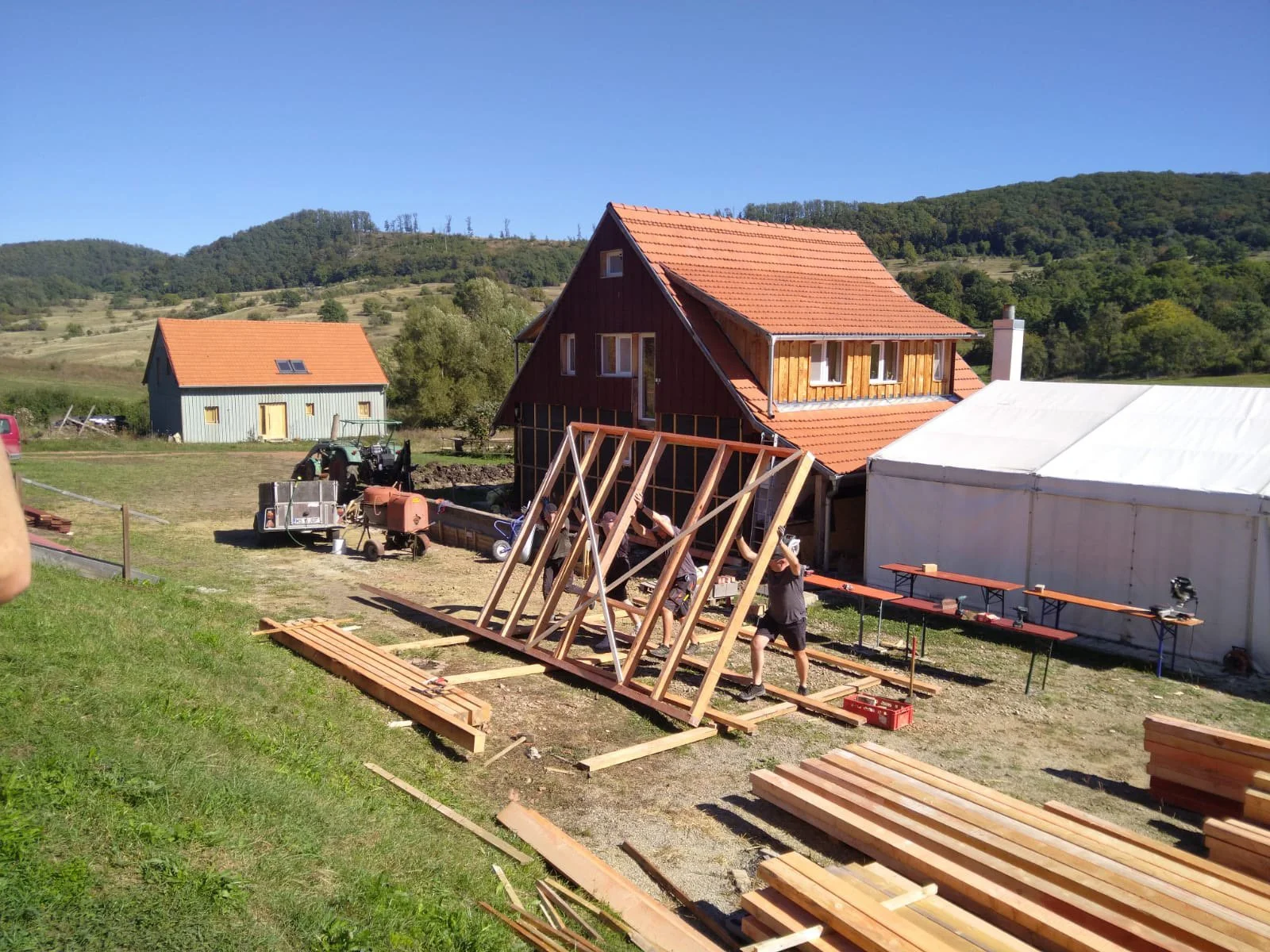 Person baut Holzrahmen für ein Zelt oder eine kleine Struktur auf einem ländlichen Grundstück bei sonnigem Wetter.