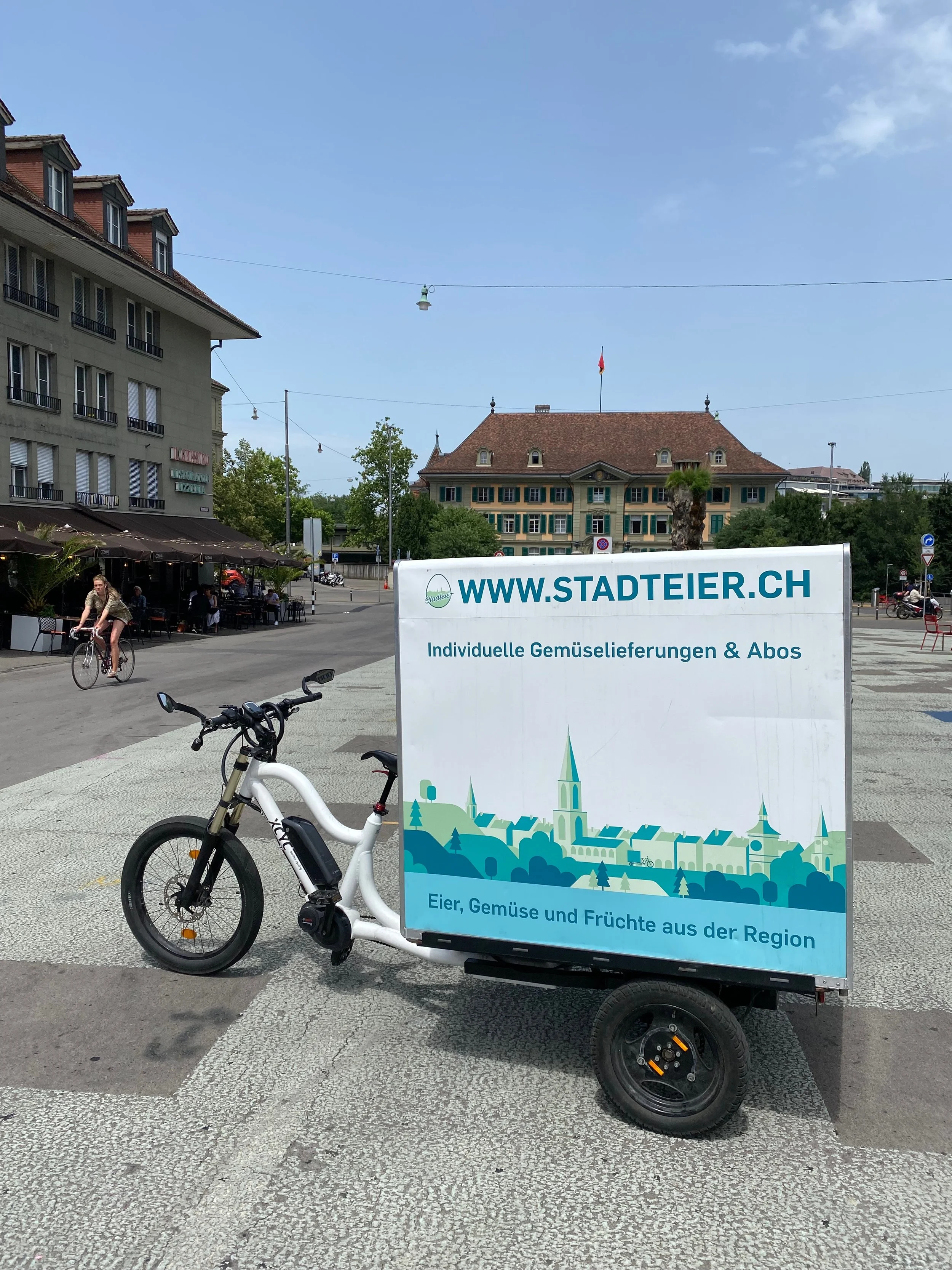 Ein mobiler Stand für regionale Eier, Gemüse und Früchte mit einem Fahrrad, steht auf einem Platz vor einem historischen Gebäude in einer Stadt. Das Schild zeigt die Webseite www.stadtteier.ch und bewirbt individuelle Gemüsellieferungen und Abos.
