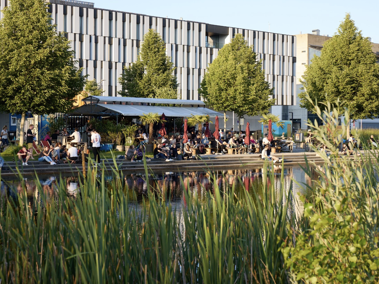 Menschen sitzen und laufen an einem Flussufer mit Bäumen und einem modernen Gebäude im Hintergrund, einige unter Sonnenschirmen, bei sonnigem Wetter.