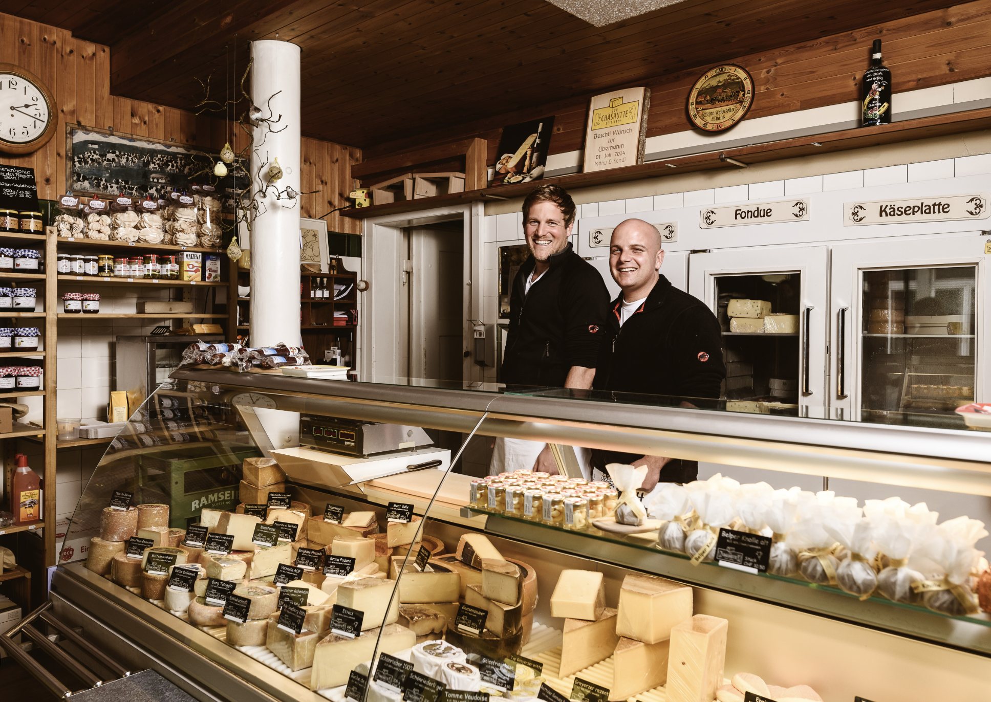 Zwei Männer hinter einem Käse- und Wursttheke in einem Käseladen, lächeln in die Kamera. Es gibt Käse, Wurst und andere Produkte im Verkaufsbereich.