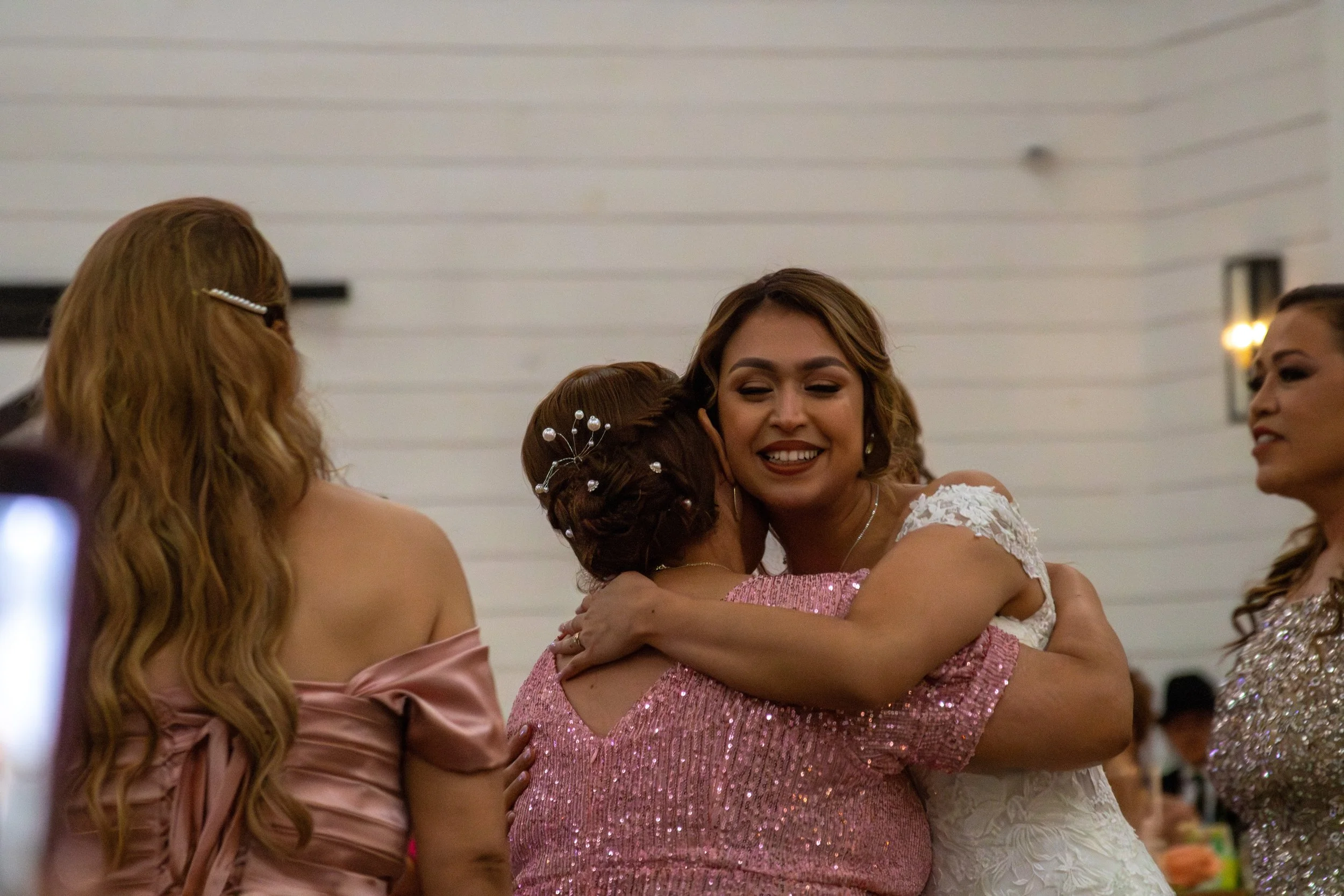 A bride and her mother embrace each other, smiling, at a celebration. The bride is on the right, wearing a white dress with lace details.