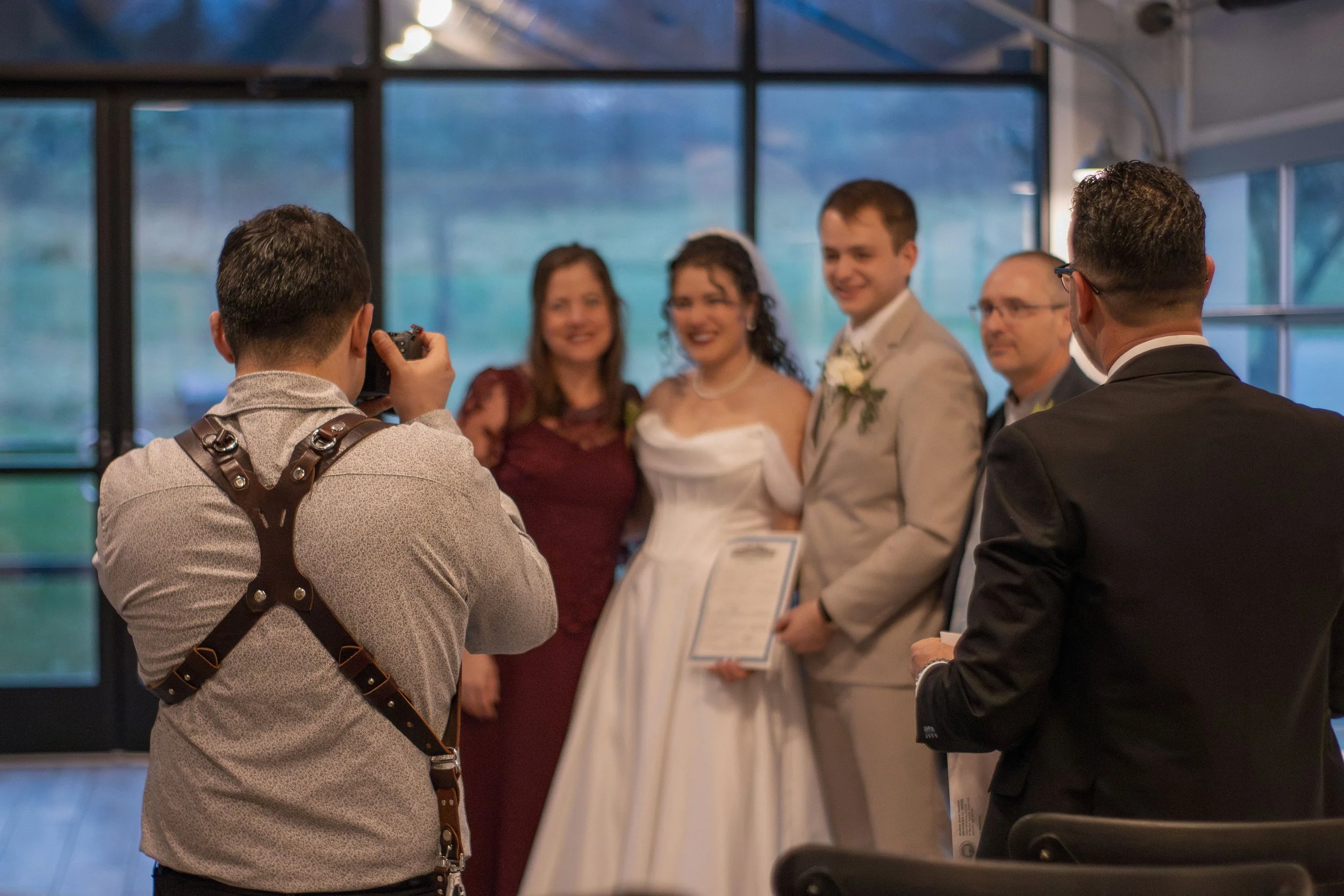 People taking a group photo at a wedding ceremony inside a building with large windows in the background.