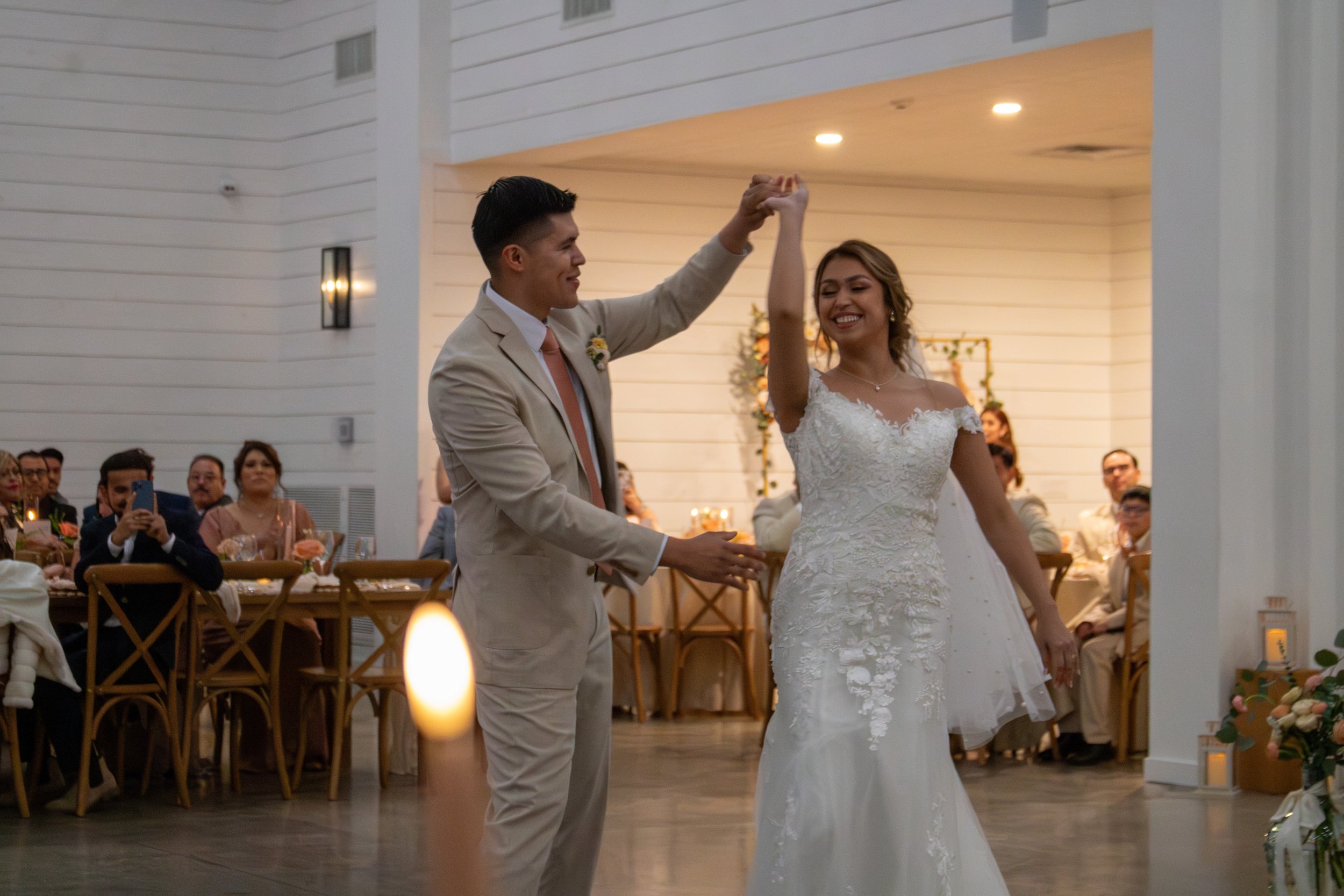 A newlywed couple dancing, with the groom spinning the bride in an indoor wedding reception. Guests are seated at tables in the background.