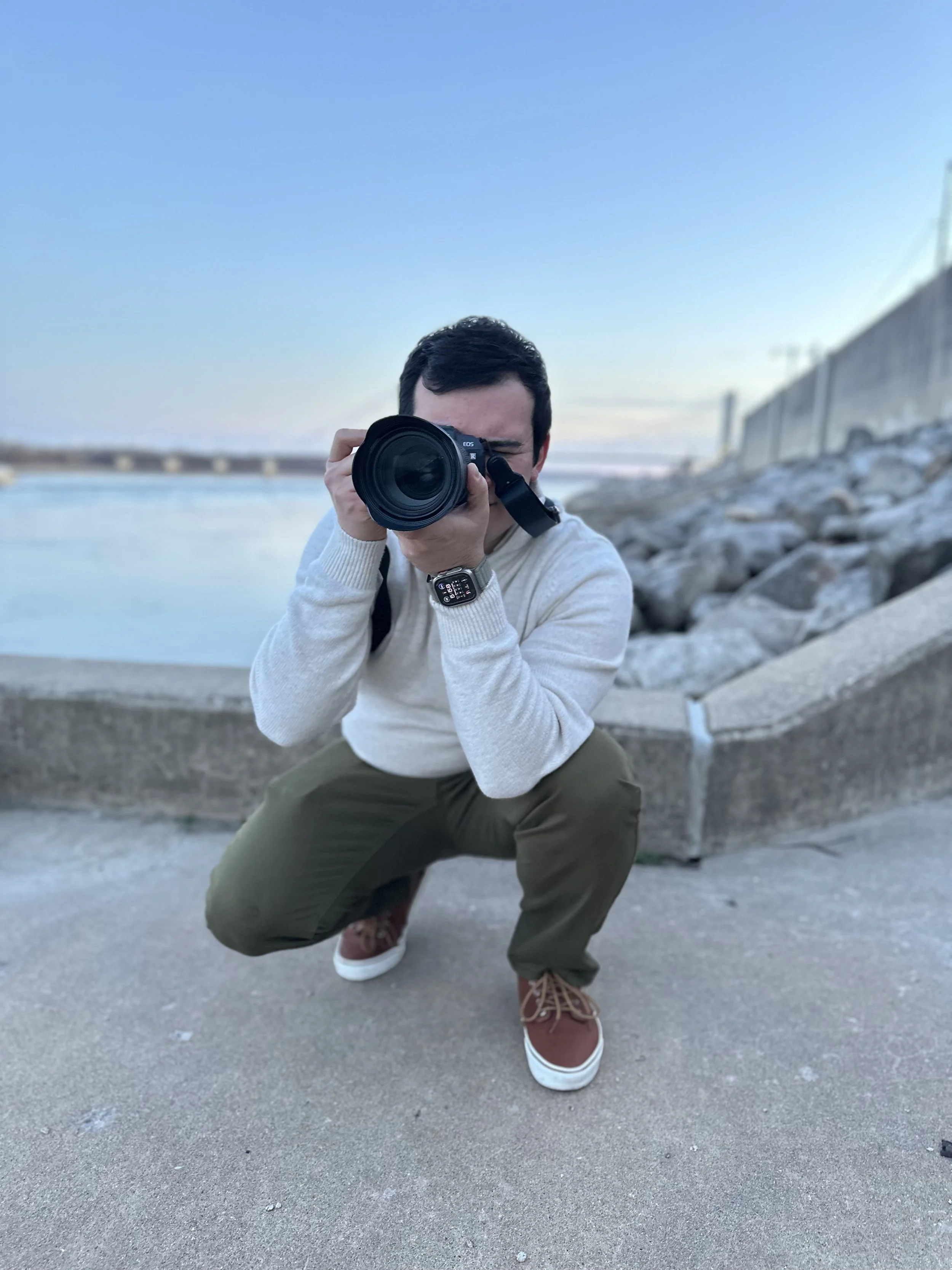 A person crouching on a concrete surface, taking a photo with a camera, outdoors near a body of water, with a rocky shoreline and a wall in the background under a clear sky.