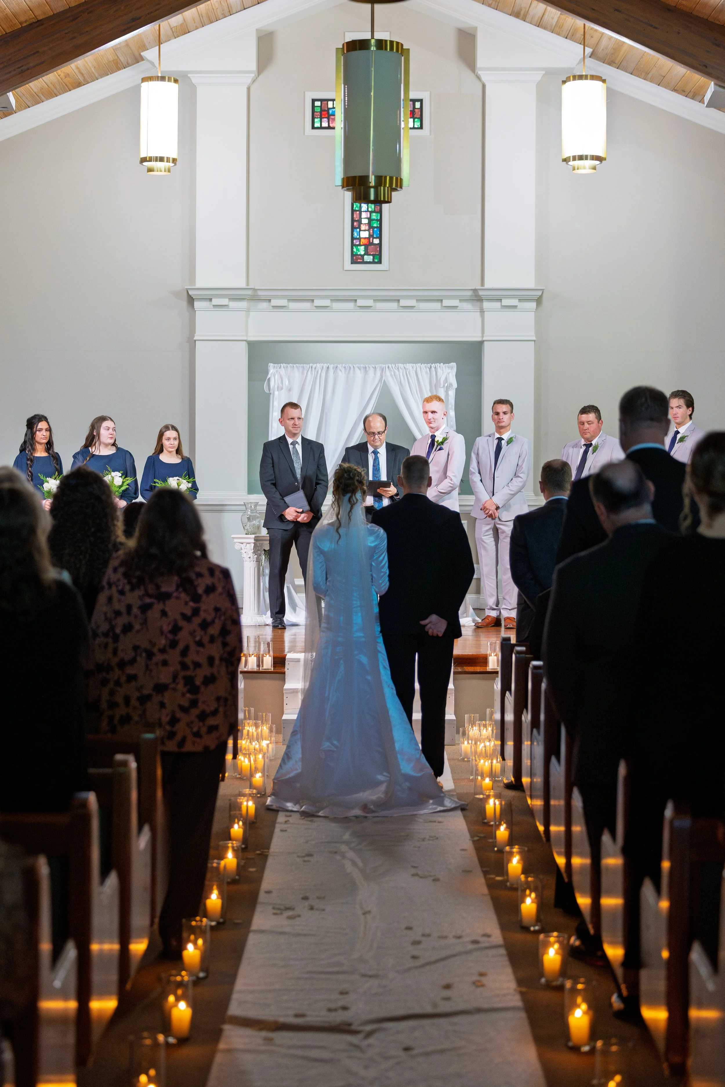 A wedding ceremony inside a church with a bride and groom standing at the altar, surrounded by guests, lit by candles along the aisle, with the officiant and bridal party present.