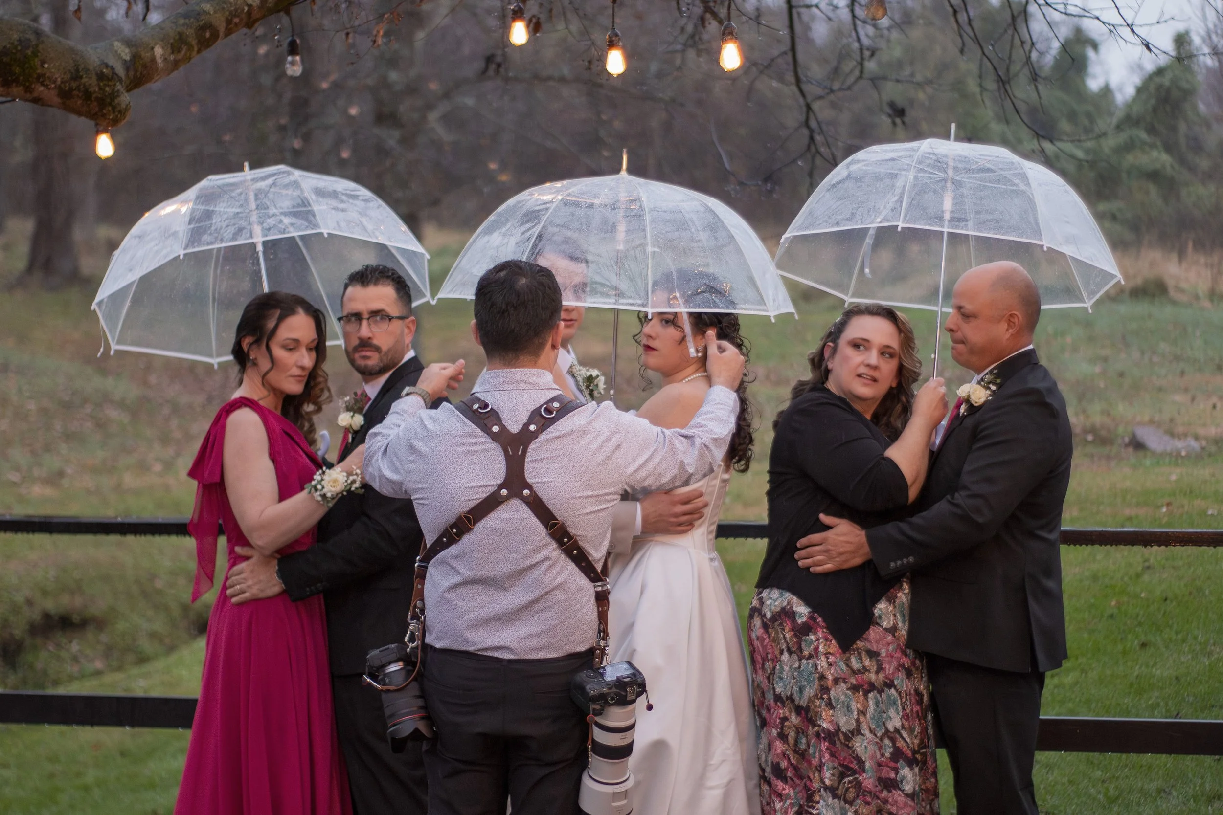 A group of seven people, including a bride in a white wedding dress, gather under umbrellas in the rain outdoors during a wedding ceremony, with greenery and string lights visible in the background.