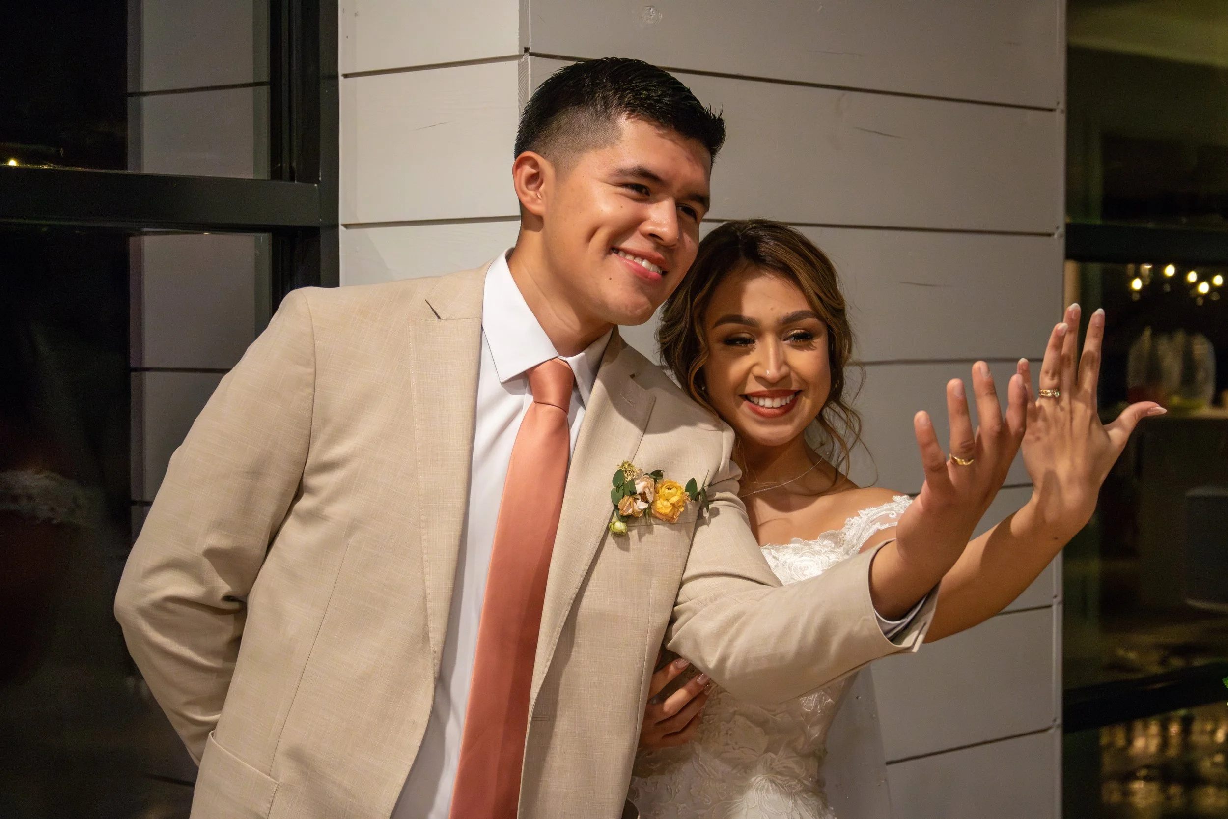 A bride and groom smiling and showing their wedding rings at a wedding reception.