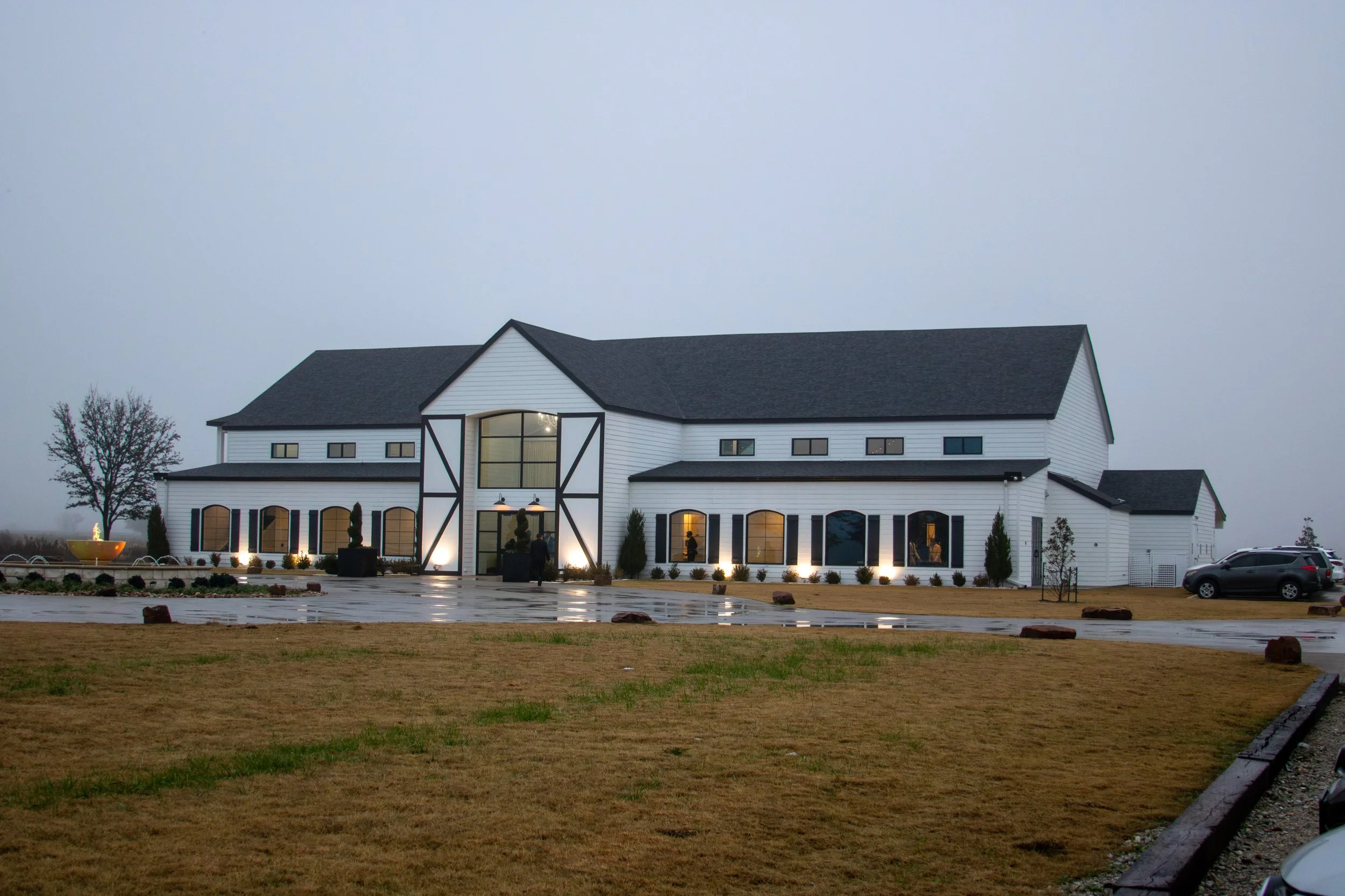 Large white barn-style building with black trim and multiple windows, lit illuminated at night, with a parking lot and grass in the foreground, cloudy sky.