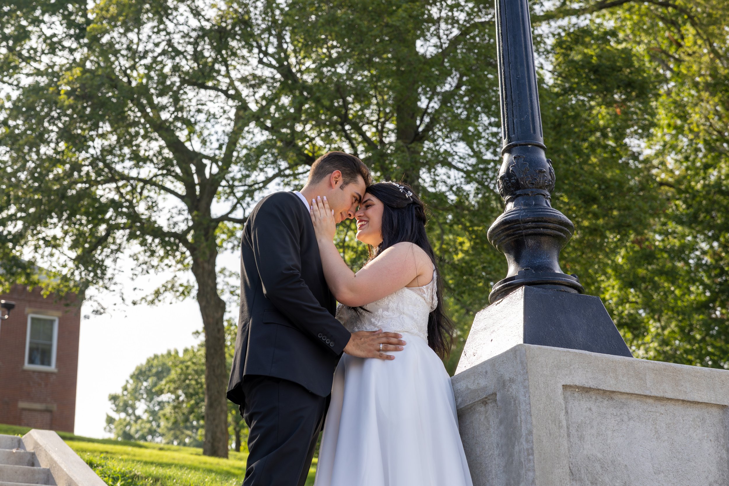 A newlywed couple sharing an intimate moment outdoors, touching foreheads and smiling, with trees and a brick building in the background.