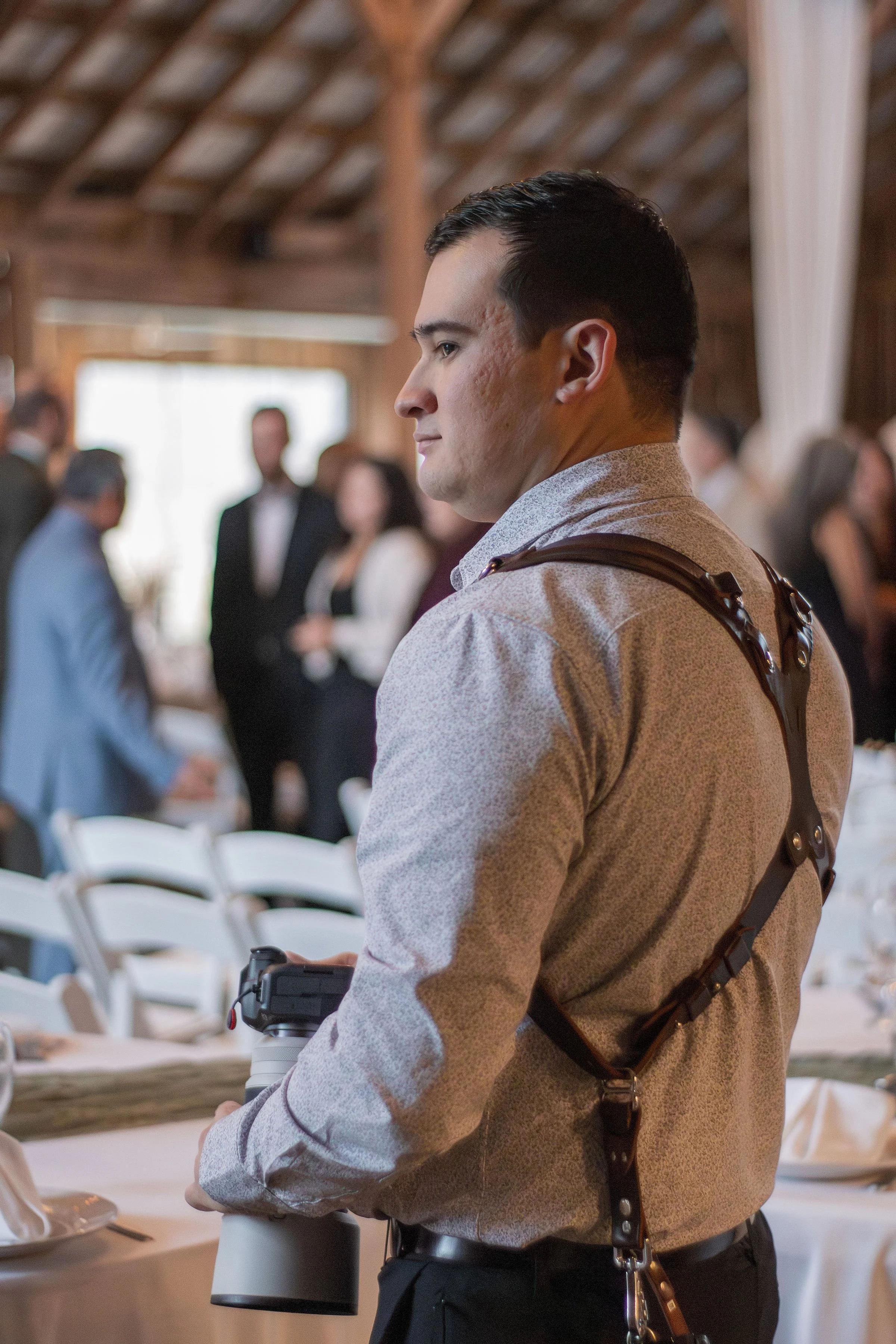 A man wearing a harness and holding a camera at a social event with people in formal attire in a rustic wooden hall.