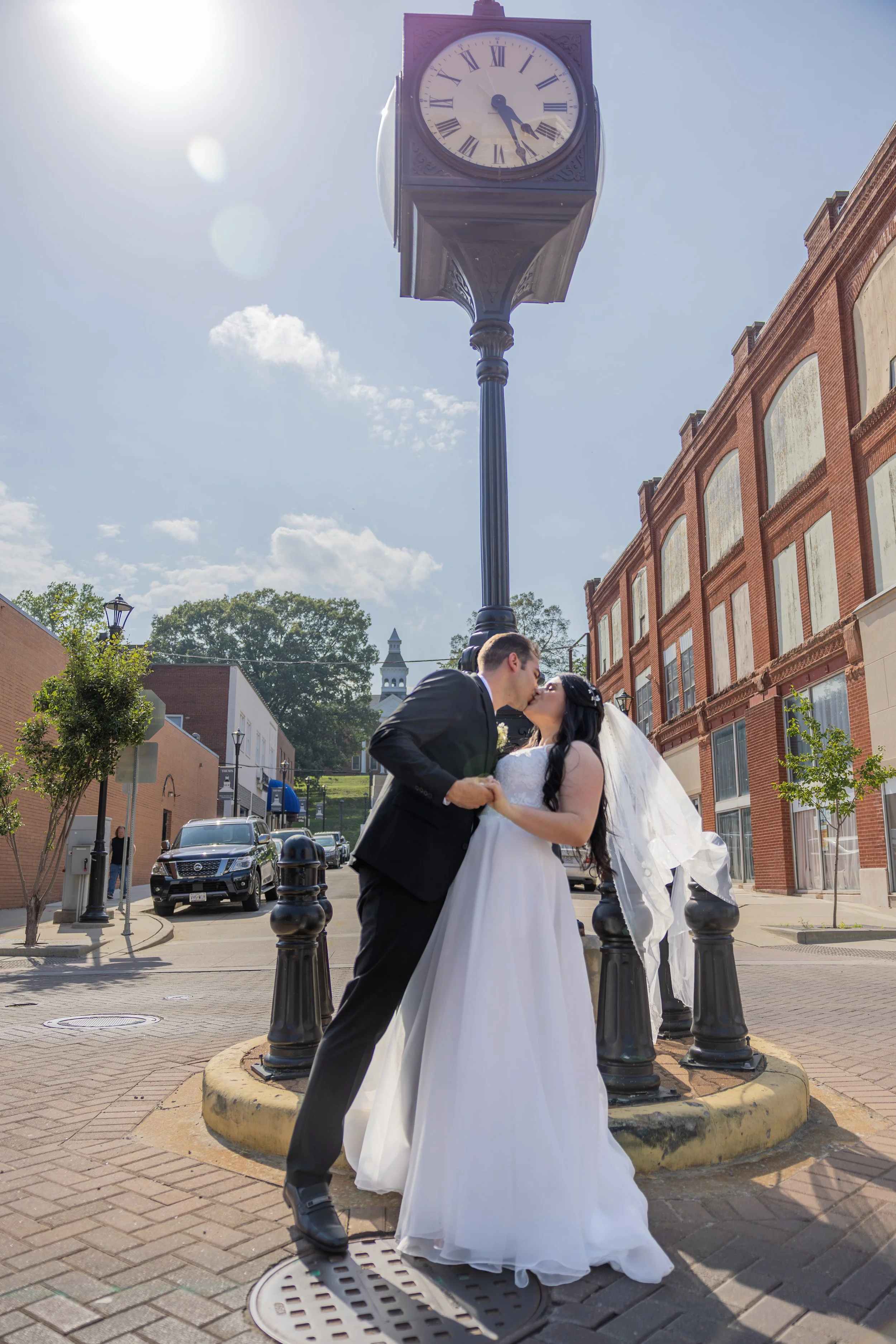 A newlywed couple sharing a kiss in front of a vintage clock on a street in a small town, with brick and white buildings and parked cars in the background.