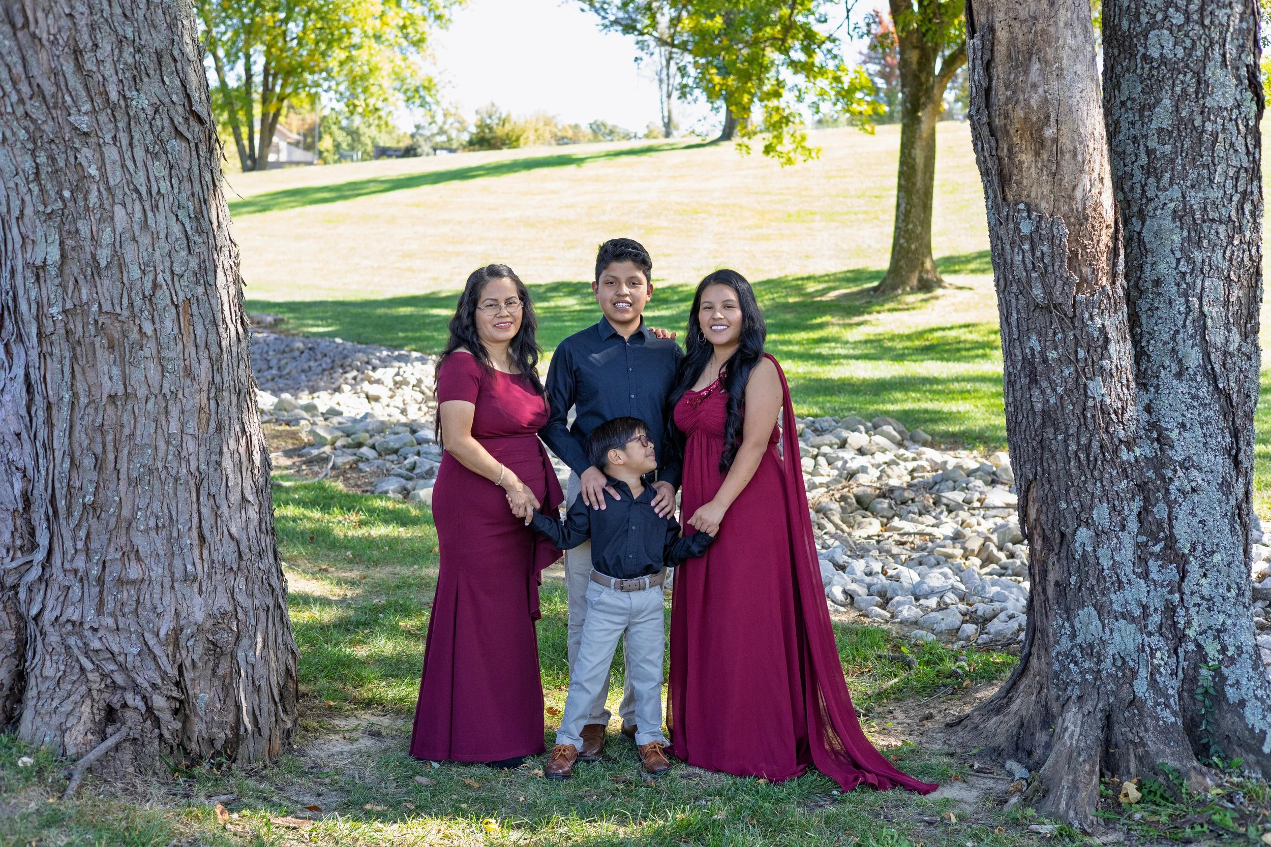 A family of four standing outdoors between two large trees on a grassy area, with a gently sloping hill and more trees in the background on a sunny day.