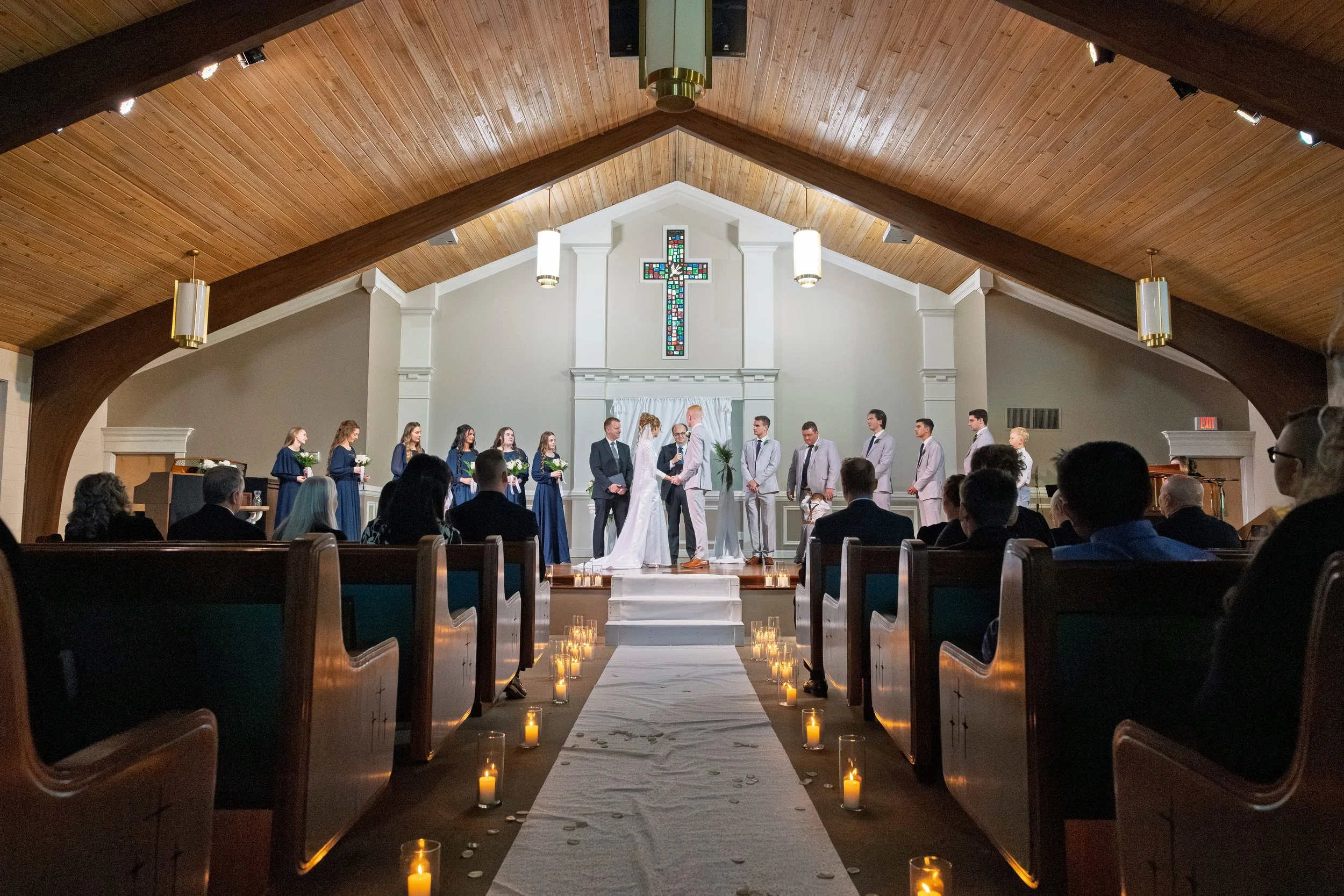A wedding ceremony taking place inside a church with a wooden ceiling, stained glass cross, and rows of pews filled with guests. The bride and groom stand at the altar with officiant and bridal party members.