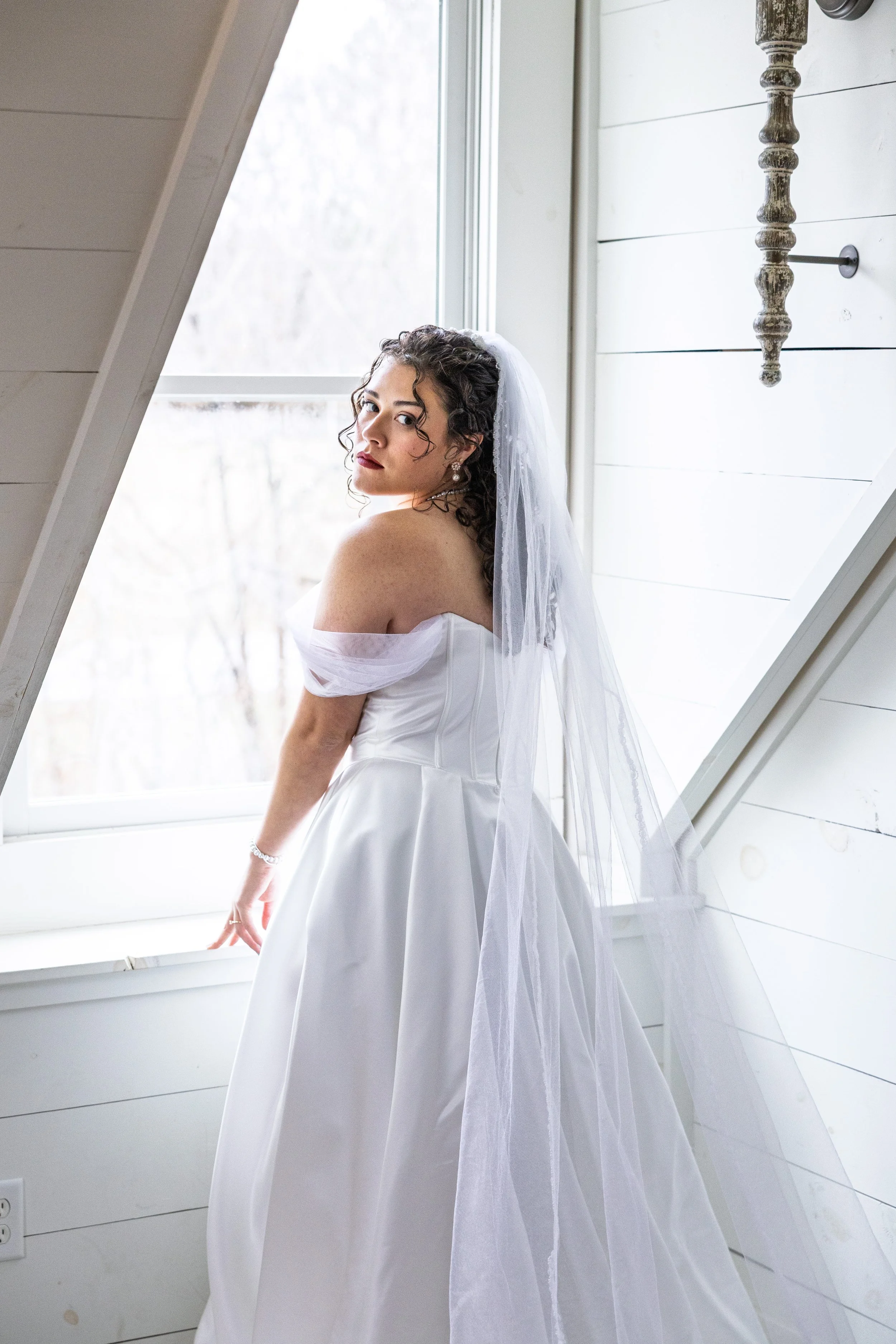 A woman in a white wedding dress and veil standing by a window in a bright room with white wooden walls.