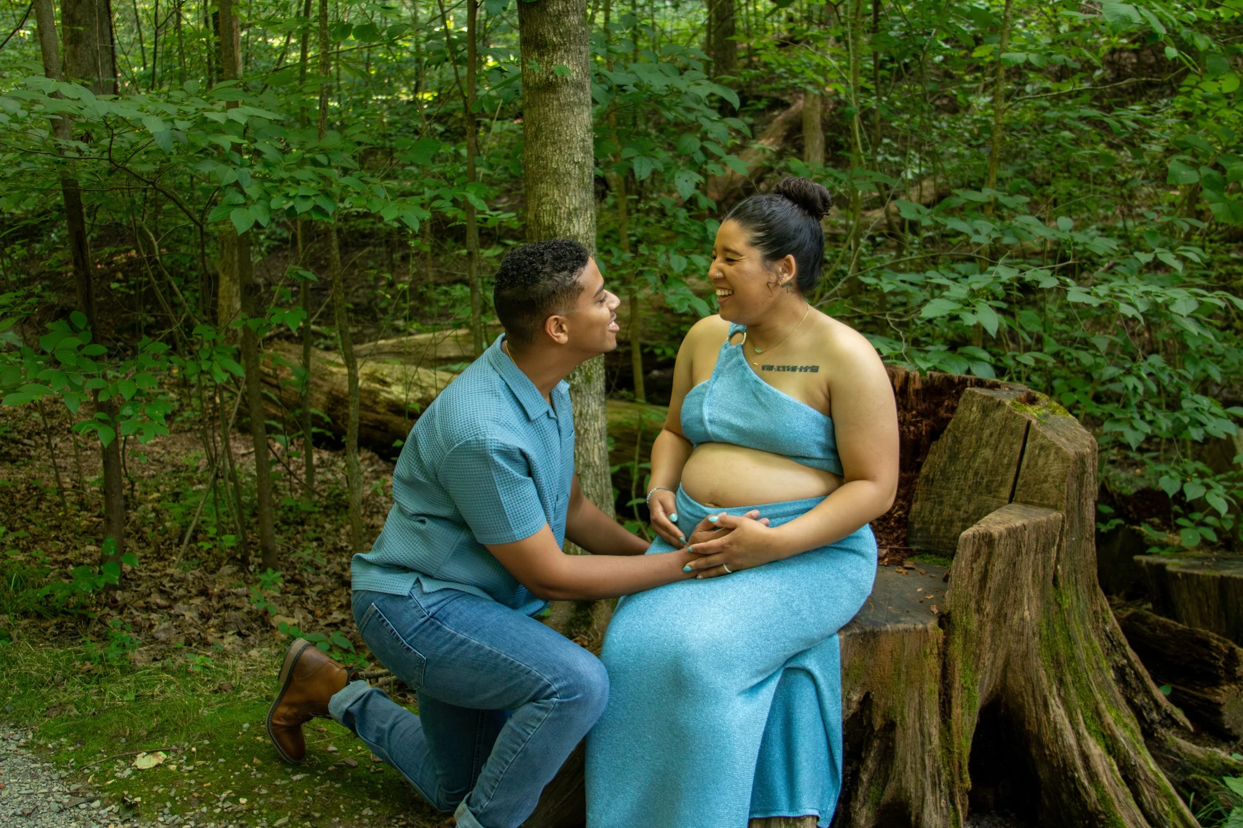 A man kneeling and holding the pregnant belly of a woman sitting on a tree stump in a lush green forest, both smiling at each other.