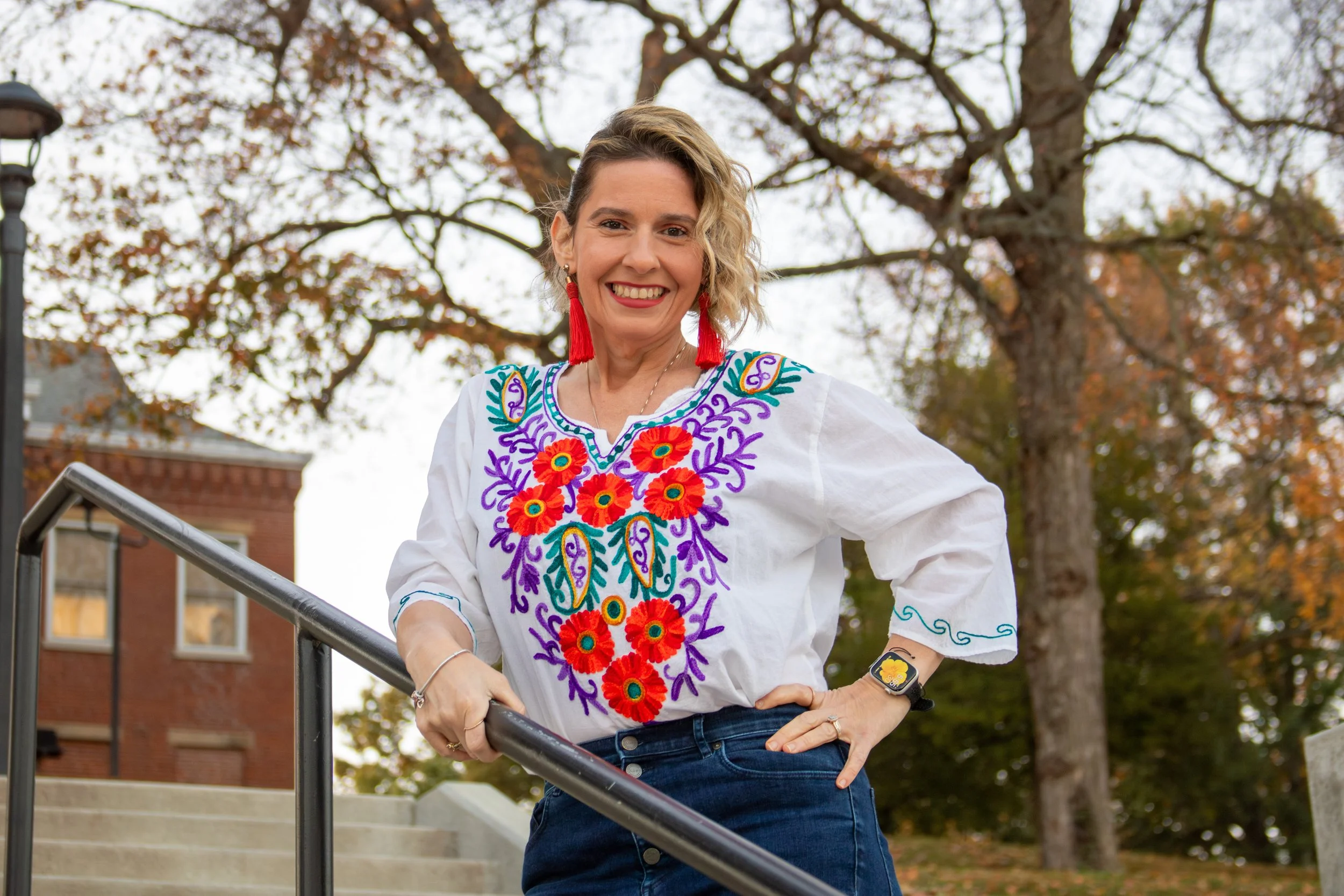Woman in a white embroidered blouse with red, purple, and green floral designs, red tassel earrings, standing outdoors on steps with trees and a brick building behind her.