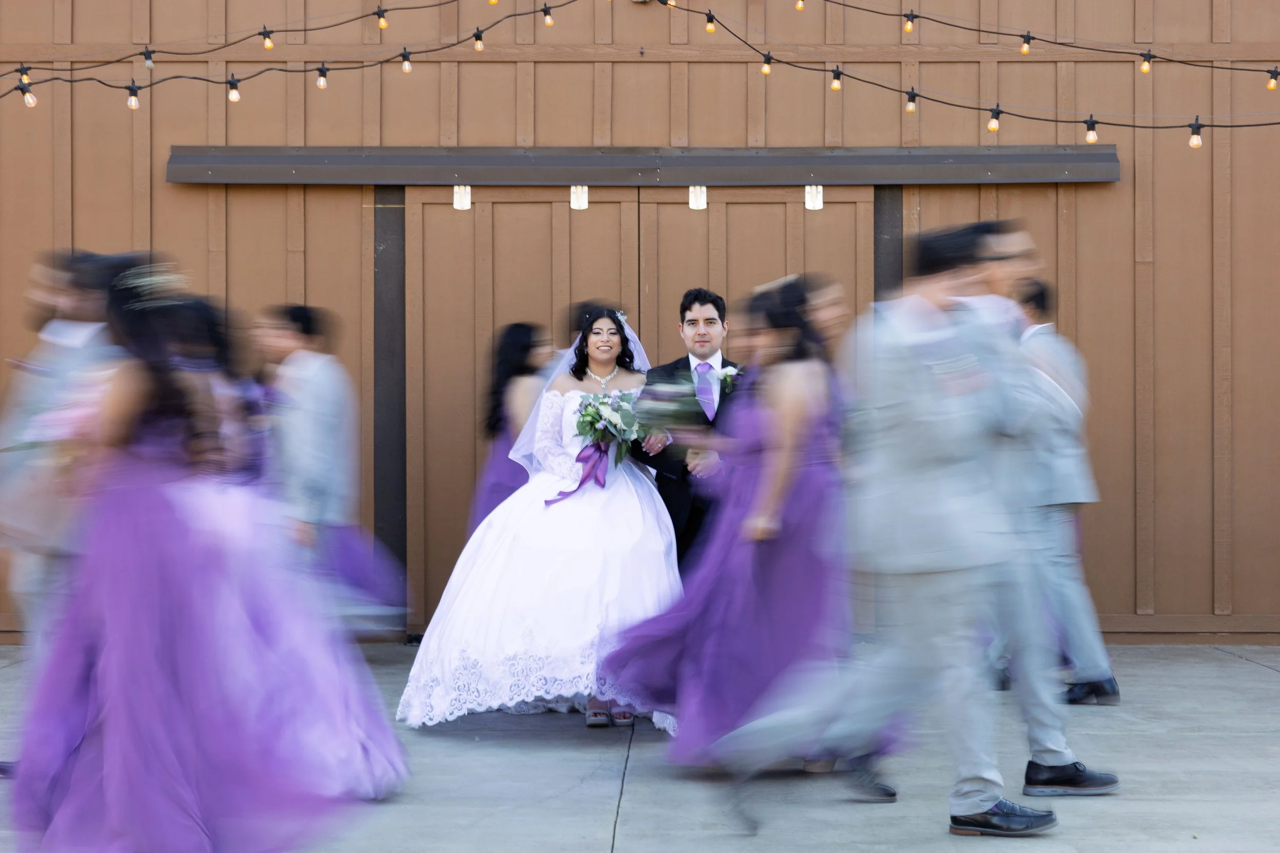 A bride in a white wedding dress holding a bouquet of flowers and a groom in a suit standing against a wooden wall with string lights above, while wedding guests in purple dresses and suits walk past blurry.