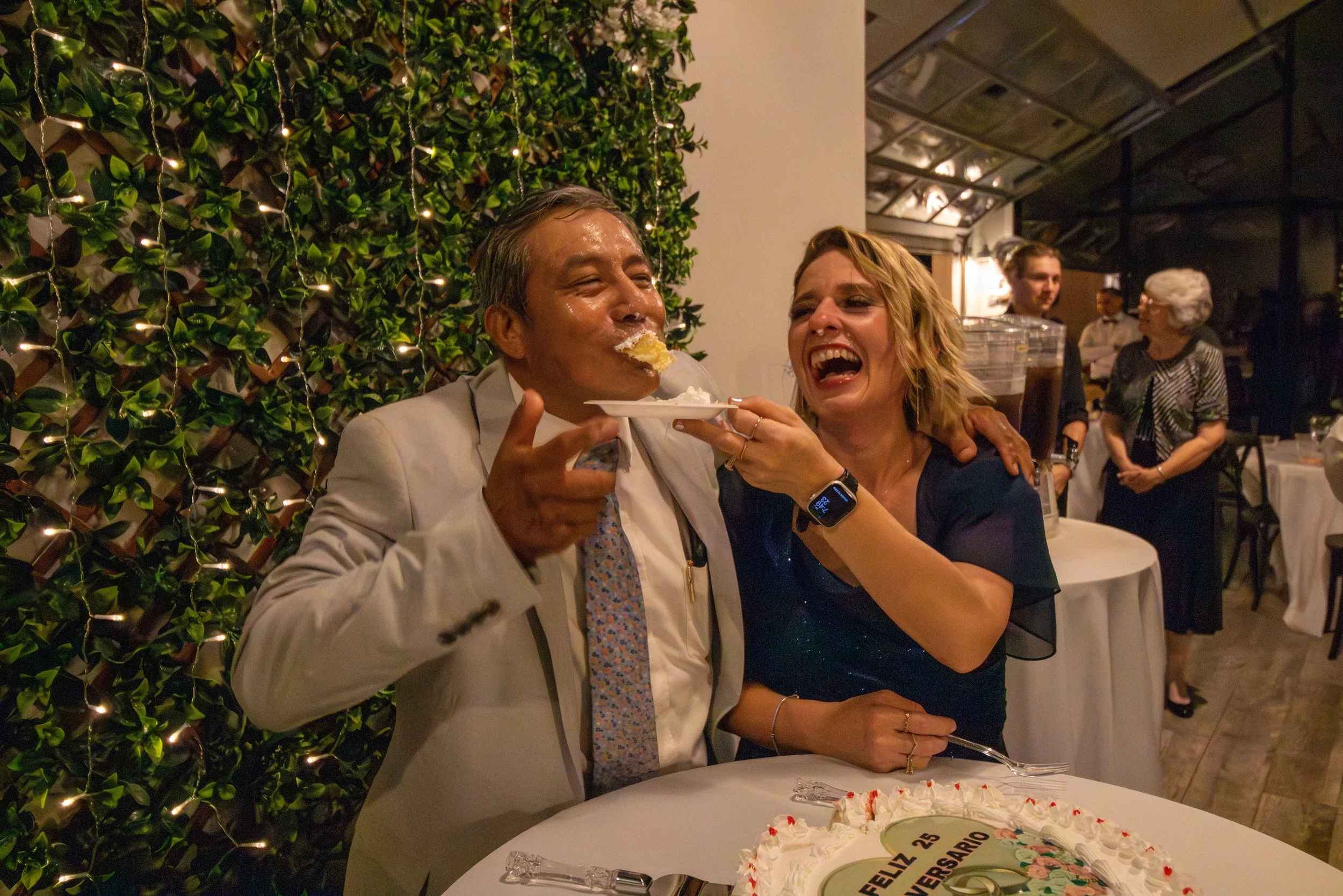 A woman is feeding a man cake at a celebration, with a cake on the table. The woman is smiling and wearing a smartwatch, and the man looks happy. There are other people in the background at a restaurant or banquet hall.
