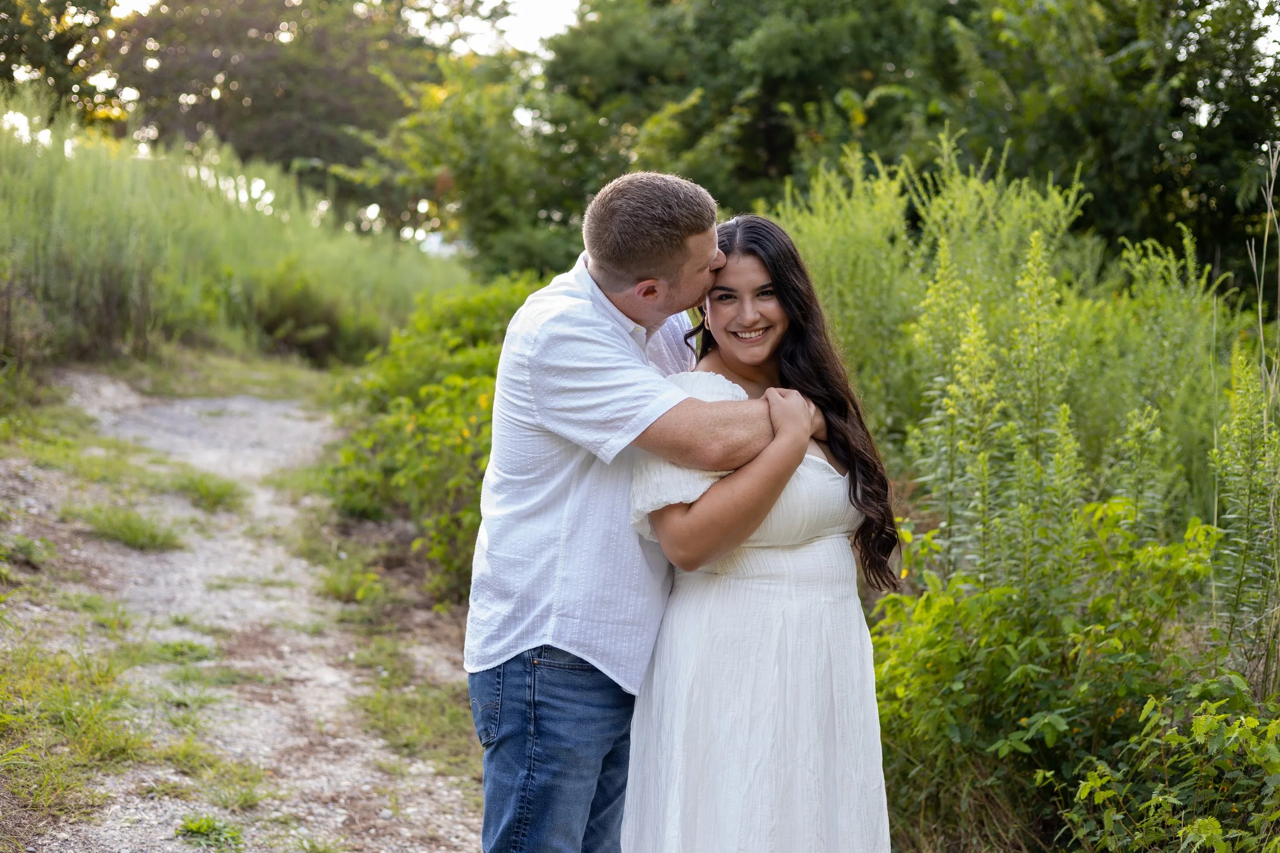 A man kisses a woman's forehead as she smiles, standing on a dirt path surrounded by greenery in a park or garden.