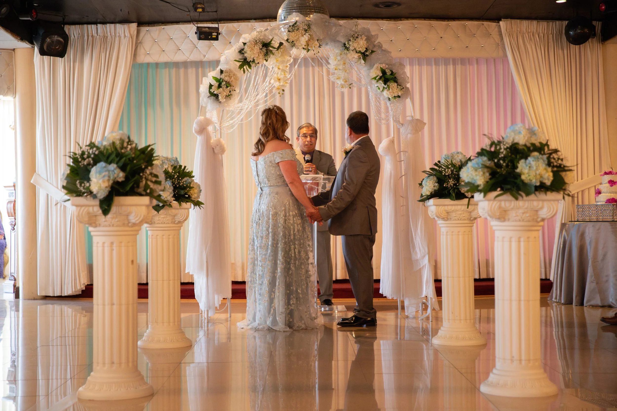A couple getting married, holding hands, standing in front of an officiant during their wedding ceremony. The ceremony takes place indoors with decorative drapery, floral arrangements, and a wedding arch adorned with white flowers and drapes.