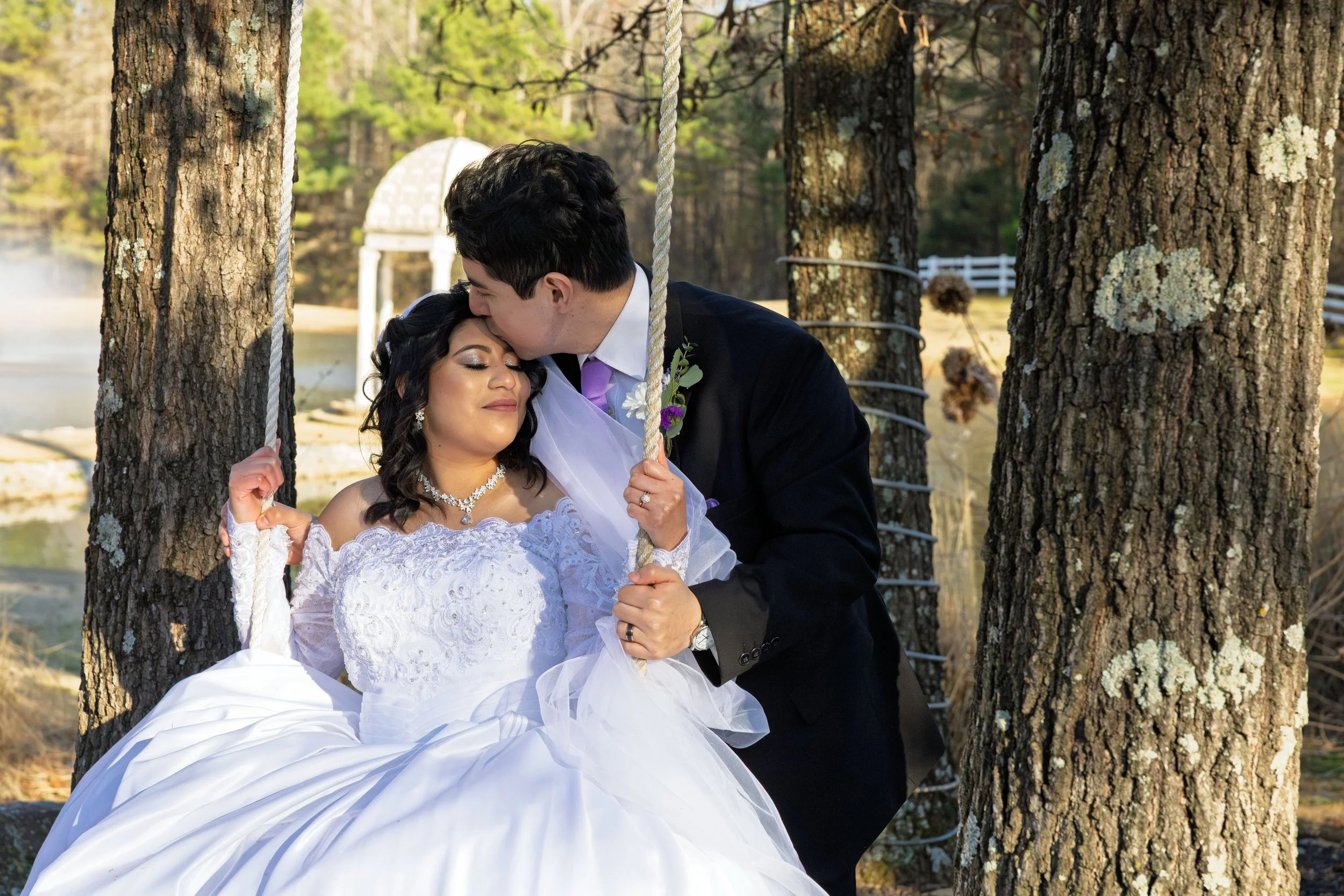 A newlywed couple in wedding attire sharing a tender moment outdoors. The bride is seated on a swing holding onto the ropes, wearing a white wedding dress with lace and jewelry, and has closed eyes with a peaceful expression. The groom is leaning in,