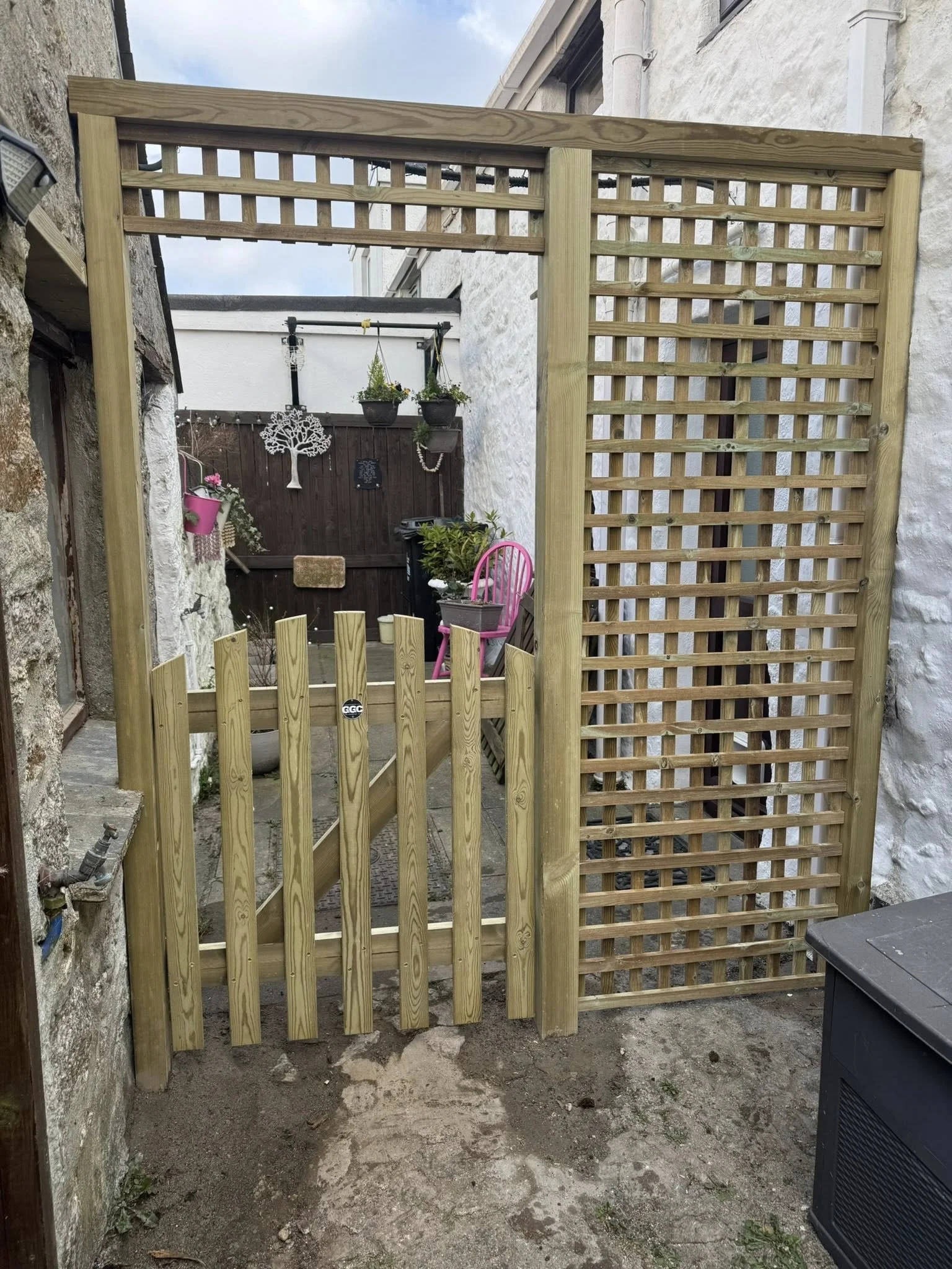 A newly constructed wooden gate with a combination of slats and lattice panels, attached to stone and white walls, blocking a small outdoor patio area.