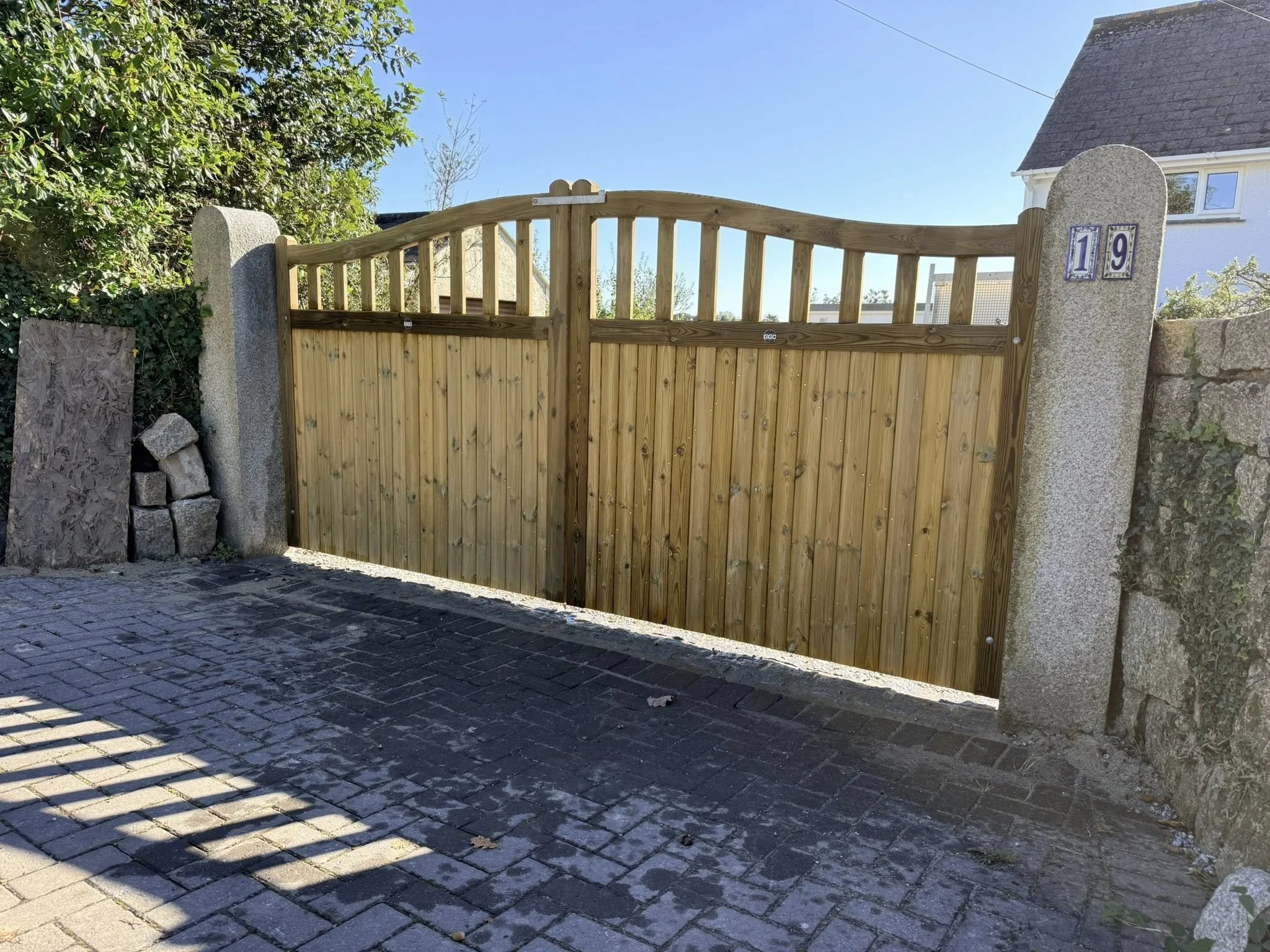 A wooden gate with decorative top, flanked by stone pillars, blocks, and a stone wall on the right side. House with white walls and a dark roof is partially visible in the background, under a clear blue sky.
