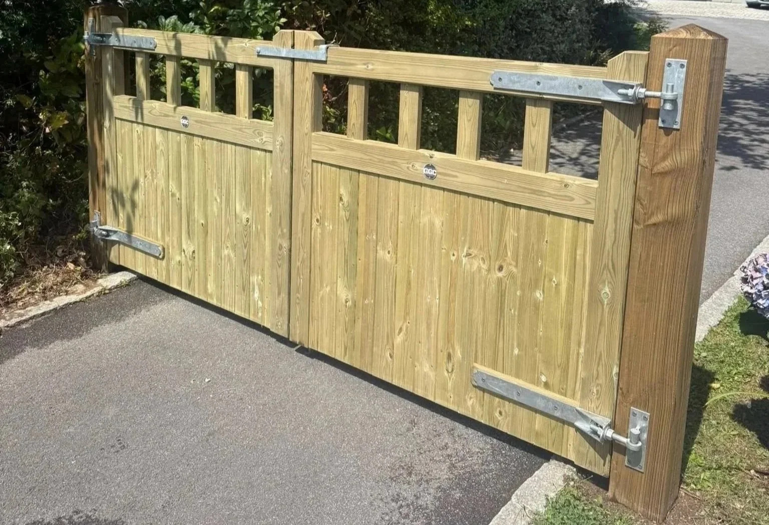 Wooden gate with metal hinges and latch, installed on a paved surface with bushes and grass nearby.