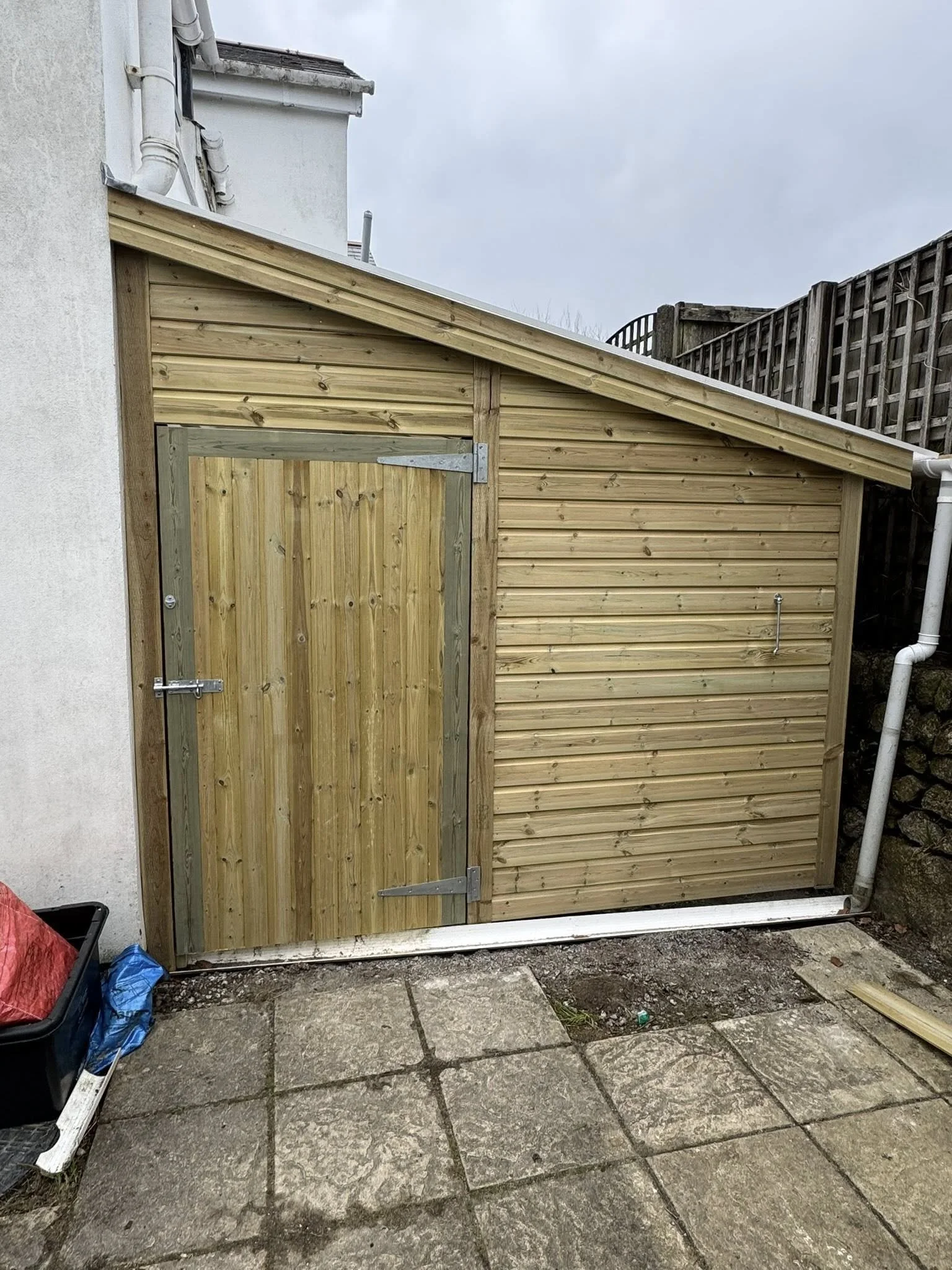 A newly built wooden shed with a sloped roof, attached to a house wall, with a small wooden door secured with metal hinges and latches, situated on a paved outdoor area.