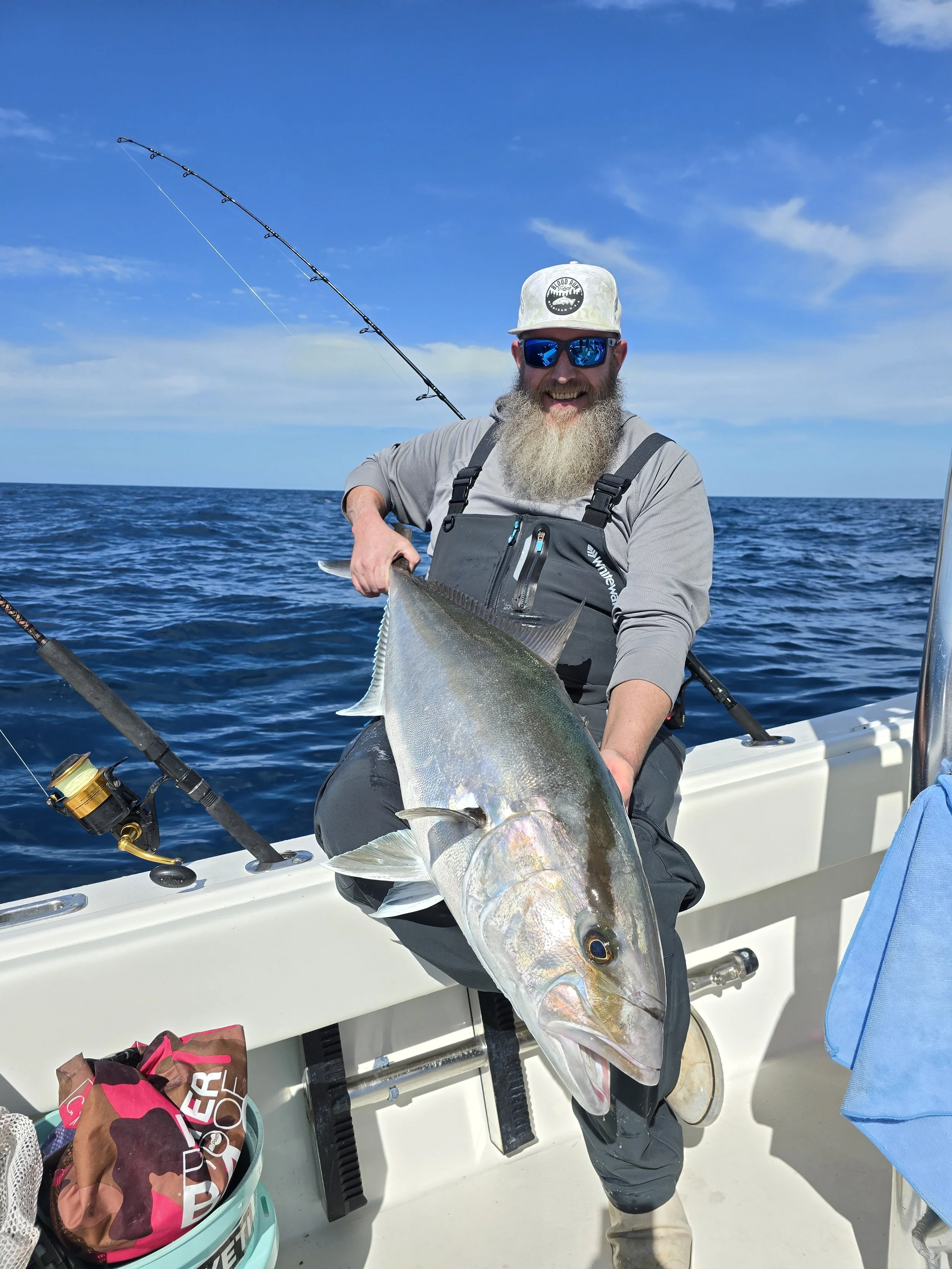 A man with a beard, wearing sunglasses, a gray cap, and fishing gear, is sitting on a boat holding a large fish, with fishing rods and ocean water in the background.