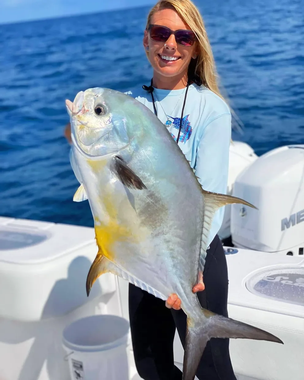 A woman on a boat holding a large yellowfin grouper fish with the ocean in the background, smiling and wearing sunglasses.